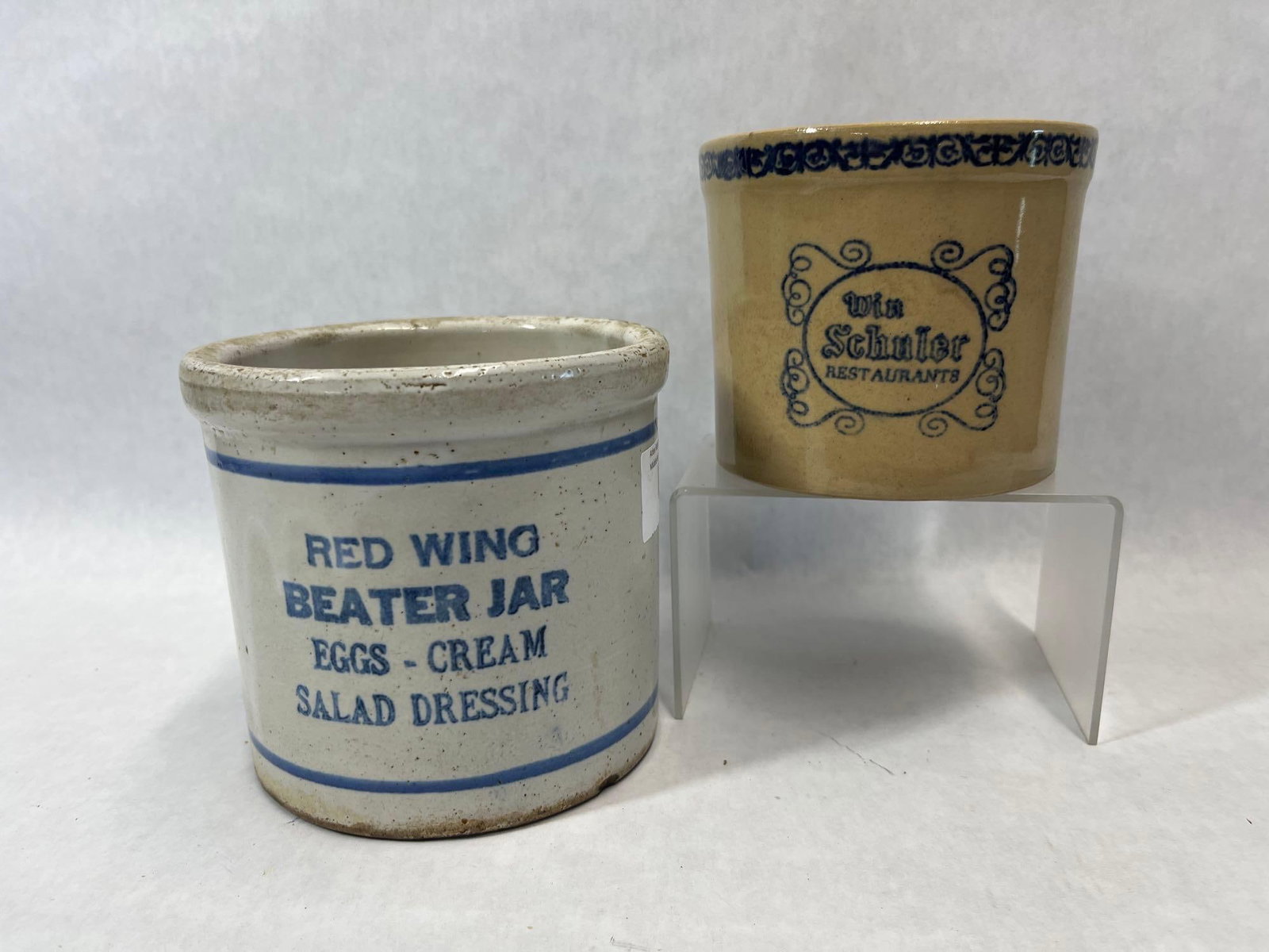Pair Red Wing & Win Schuler Advertising Crocks: This lot features two examples of 20th-century American advertising stoneware. The first item is a Red Wing Beater Jar, constructed in a cylindrical form with a reinforced, flared rim. It features a