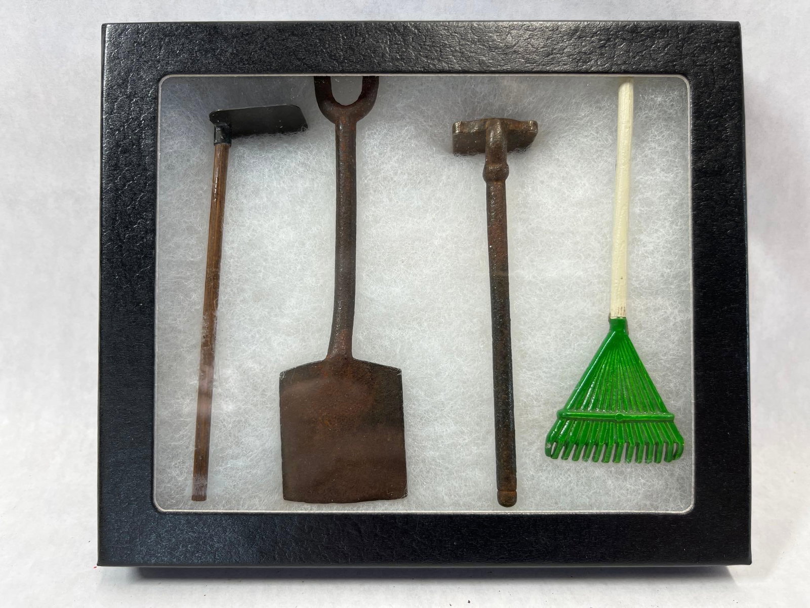 Set 4 Miniature Garden Tools in Shadow Box: A set of four miniature garden tools presented in a black-framed shadow box. The collection includes a metal spade with a D-style handle and an oxidized rusted finish, a metal tamper with a rusted