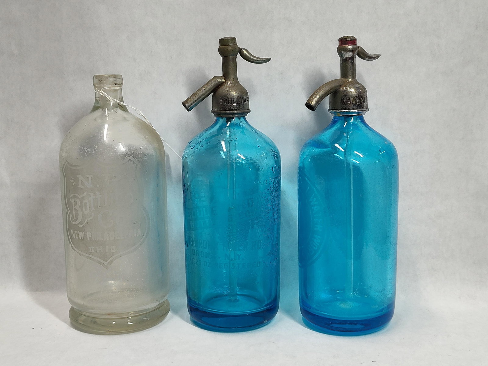 A Trio of Advertising Glass Bottles Including New York Seltzer Siphons: This lot features three glass advertising bottles from the early to mid-20th century. The collection includes two cobalt blue seltzer siphons and one clear glass bottle. The first seltzer siphon is