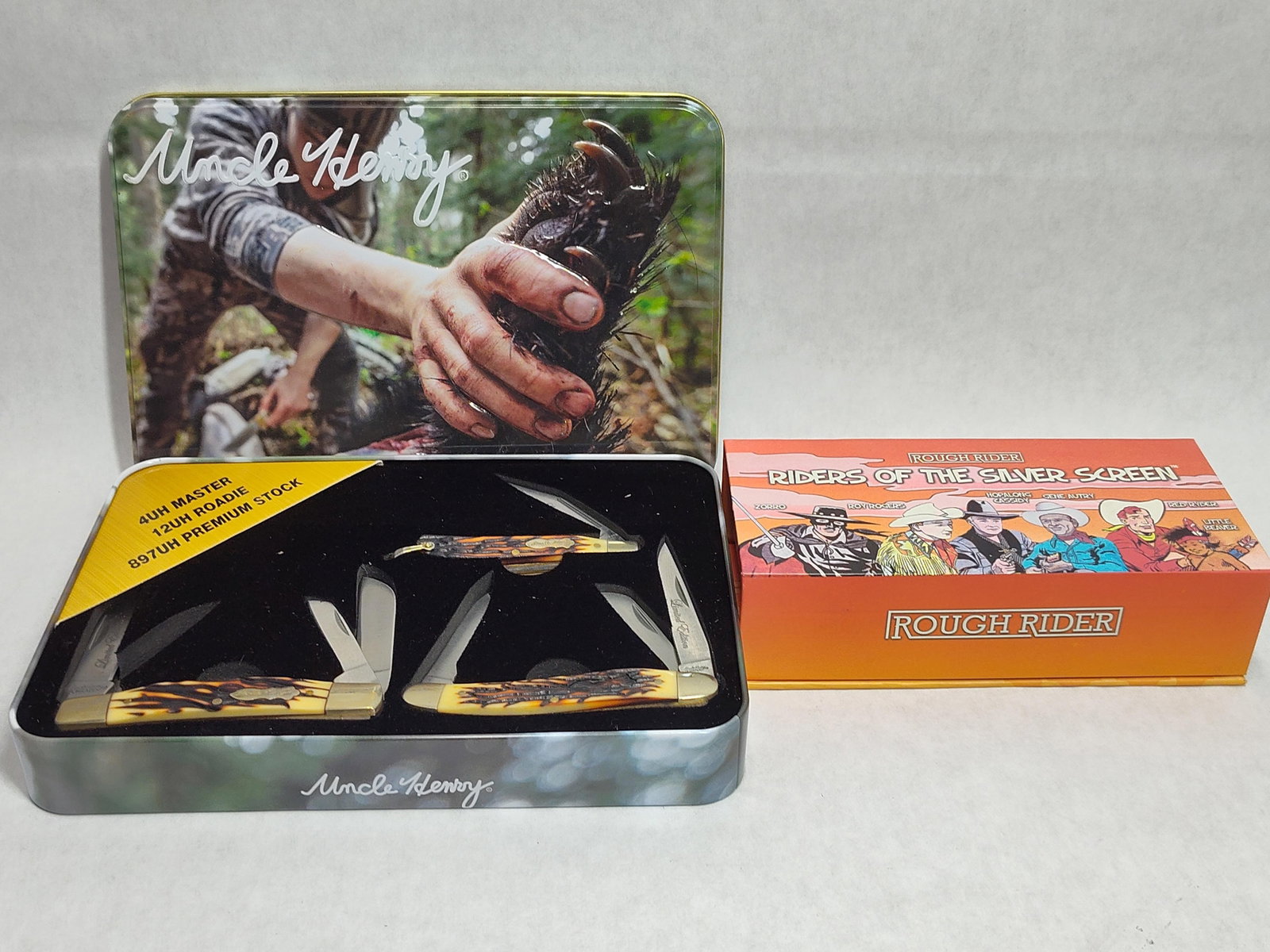 Schrade Uncle Henry 3-Knife Set & Rough Rider: This auction lot features two sets of collectible pocket knives. The first is a Schrade Uncle Henry Limited Edition gift set contained within a rectangular tin lid featuring an image of a hunter and a