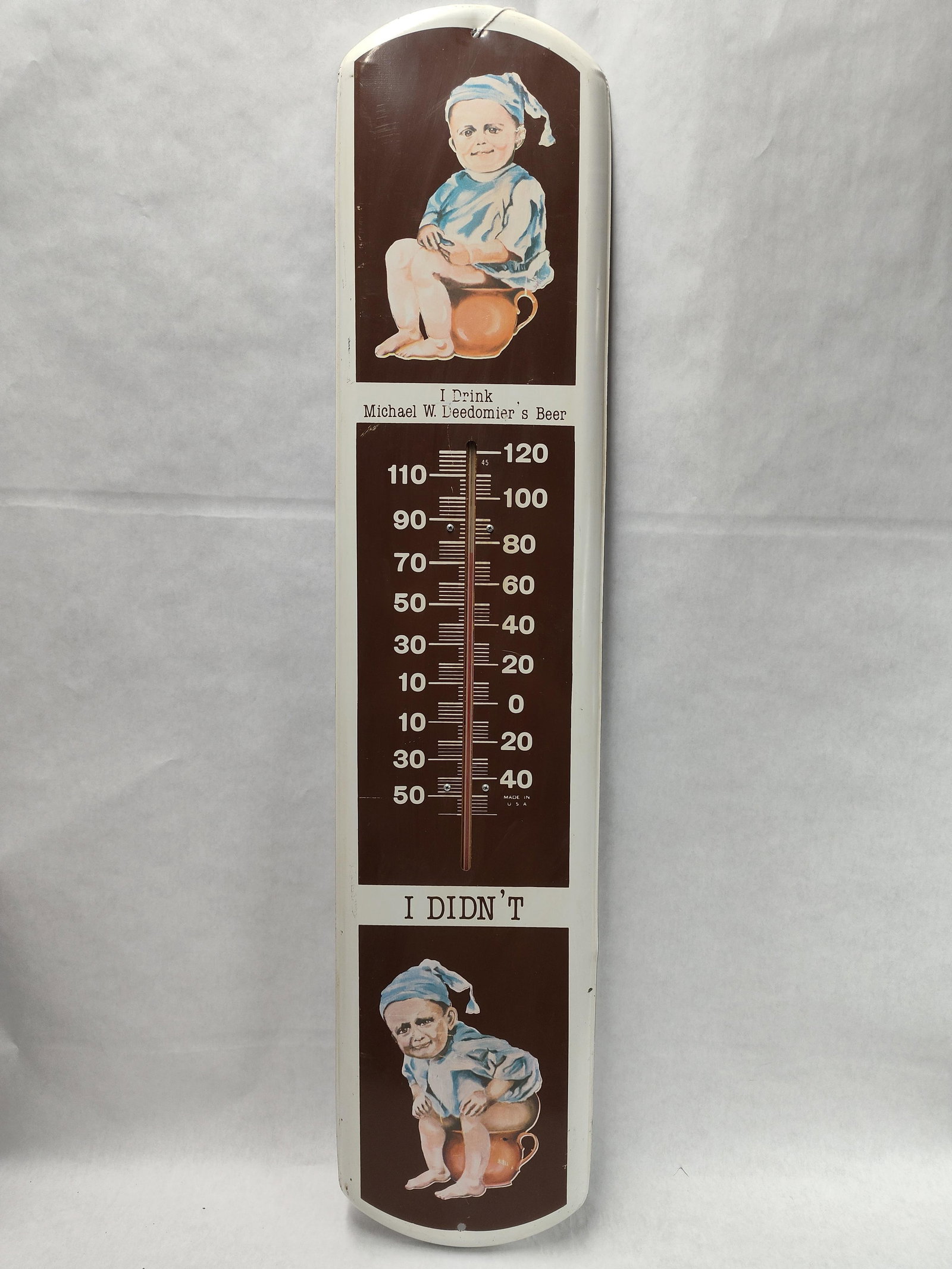 Michael W. Deedomier Beer Advertising Thermometer: This is a lithographed metal advertising thermometer featuring the Michael W. Deedomier's Beer novelty gag. The sign is designed in three vertical sections. The upper portion depicts a smiling baby