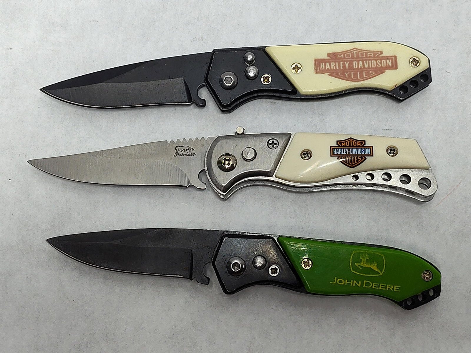 Harley-Davidson & John Deere Folding Pocket Knives: A collection of three branded folding pocket knives, attributed as automatic or spring-assisted models based on the button-lock bolster triggers and market consensus. The group includes two