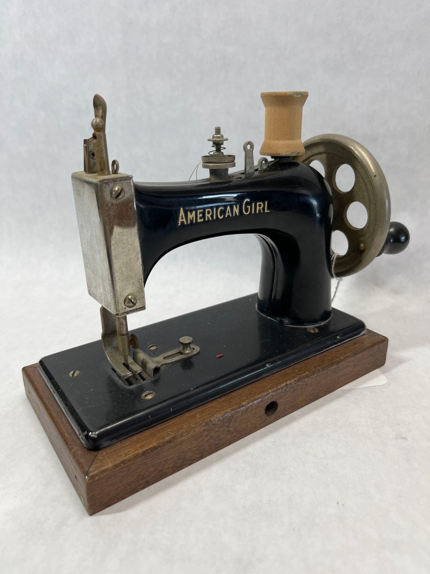 American Girl Child's Toy Sewing Machine: American Girl brand toy sewing machine, a model often associated with the 1930s. The machine is constructed of a cast metal body with a black enamel finish, mounted on a rectangular wooden base. The b