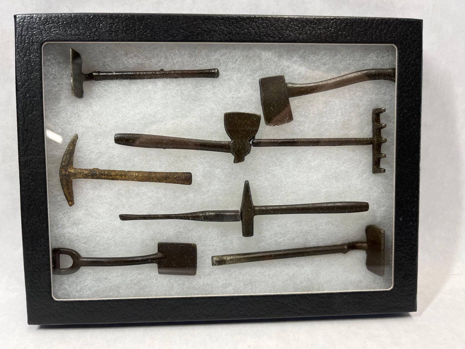 Miniature Forged Iron Hand & Garden Tool Shadowbox: A collection of miniature tools crafted from forged or cast iron with a dark, aged patina. The assortment includes various trade and garden implements such as a sledgehammer with a curved handle, a fe