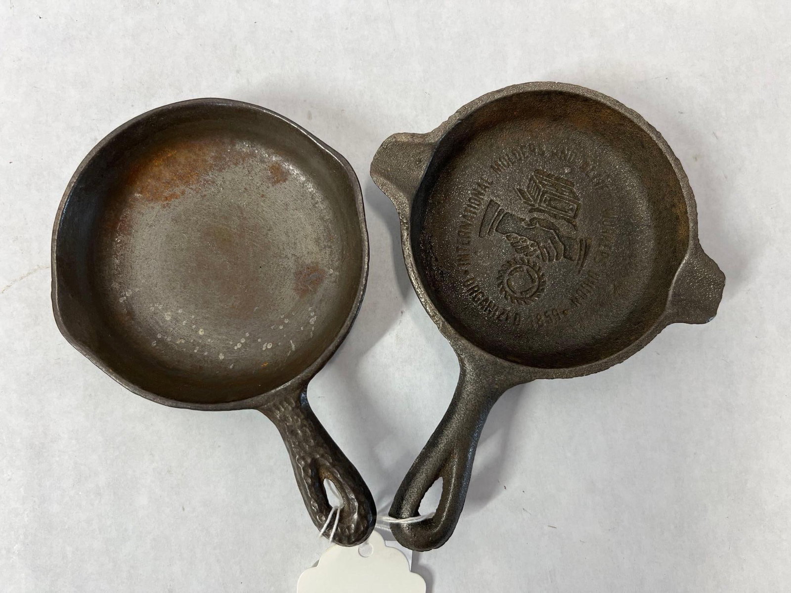 Pair Cast Iron Intl Molders & Workers Skillets: This lot comprises two miniature cast iron skillets, traditionally used as novelty ashtrays, salesman samples, or paperweights. The first item is a hammered finish skillet featuring a decorative