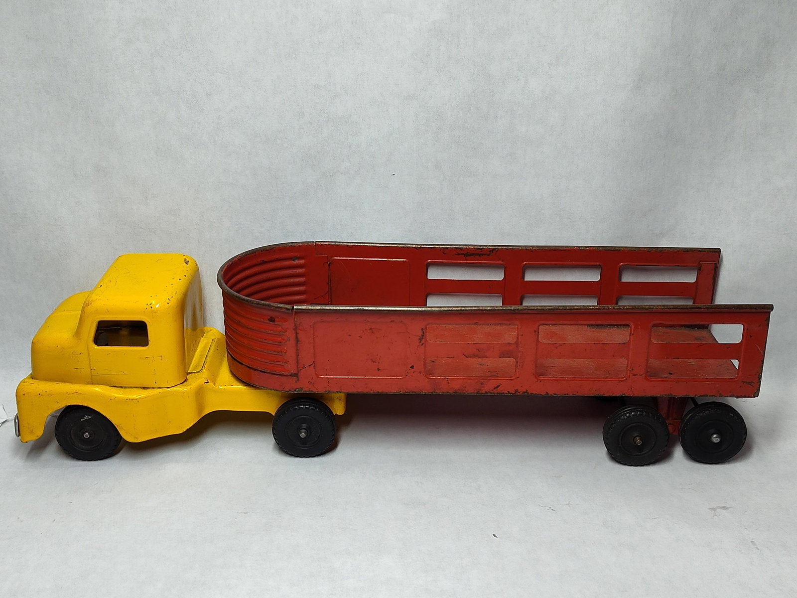 Structo Pressed Steel Truck & Open-Slat Trailer: A Structo pressed steel semi-truck and articulated open-slat trailer set. The cab is finished in yellow paint with a silver-painted front bumper. The red-painted trailer features a rounded corrugated 
