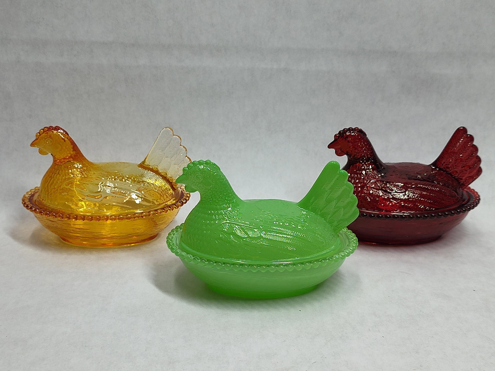 Trio Pressed Glass Hen on Nest Covered Dishes: This collection features three pressed glass figural covered dishes in the classic hen on nest form, attributed to the Indiana Glass Company. The group includes three distinct color variations: