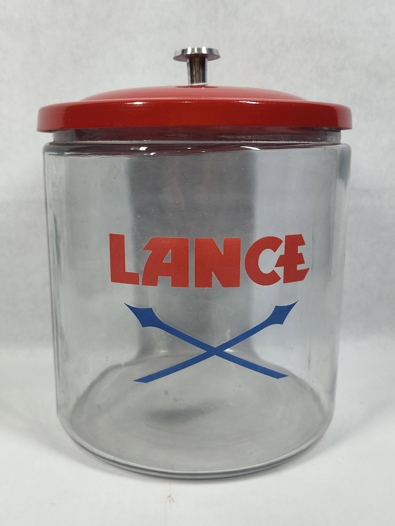 Lance Advertising Glass Jar w/ Red Metal Lid: This is a countertop snack display jar for the American brand Lance, Inc. The item features a cylindrical clear glass body with a red painted metal lid. The front of the jar is decorated with red and