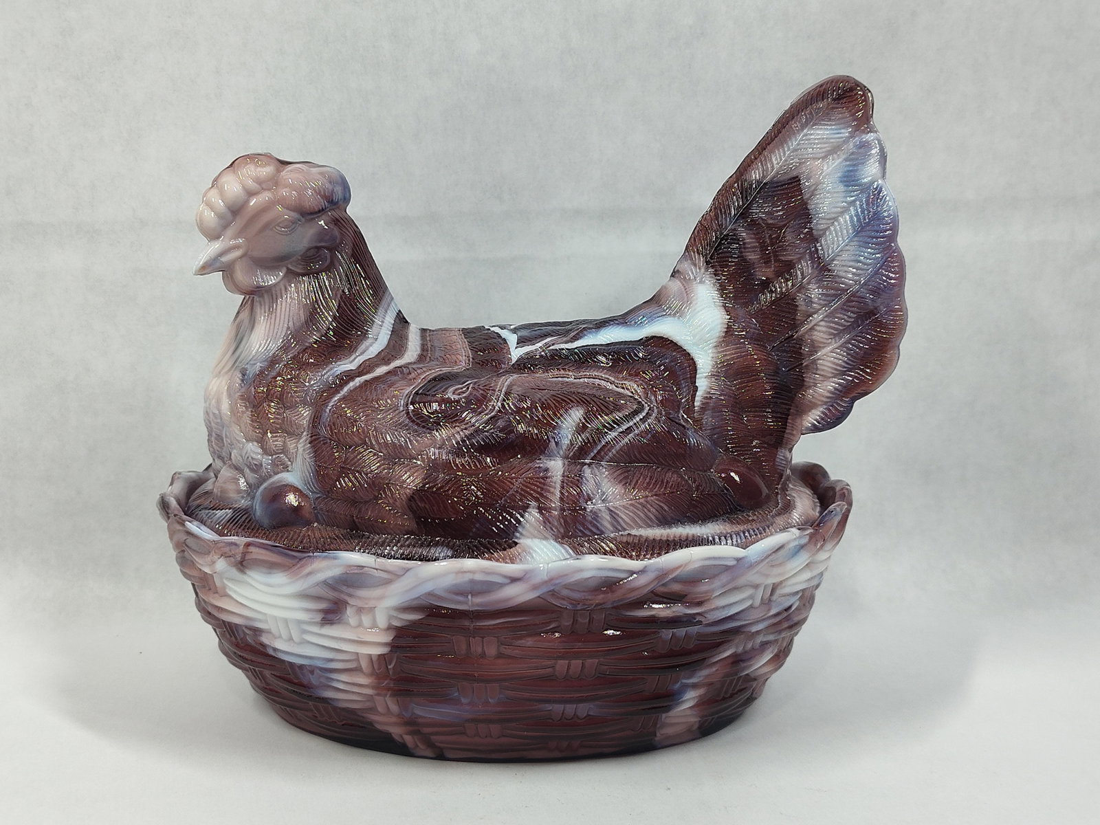 Imperial Glass Purple Slag Hen Nest Covered Dish: This covered animal dish is crafted in the form of a nesting hen resting on a basketweave base. The piece is made of purple slag glass, a type of pressed marble glass characterized by its distinctive