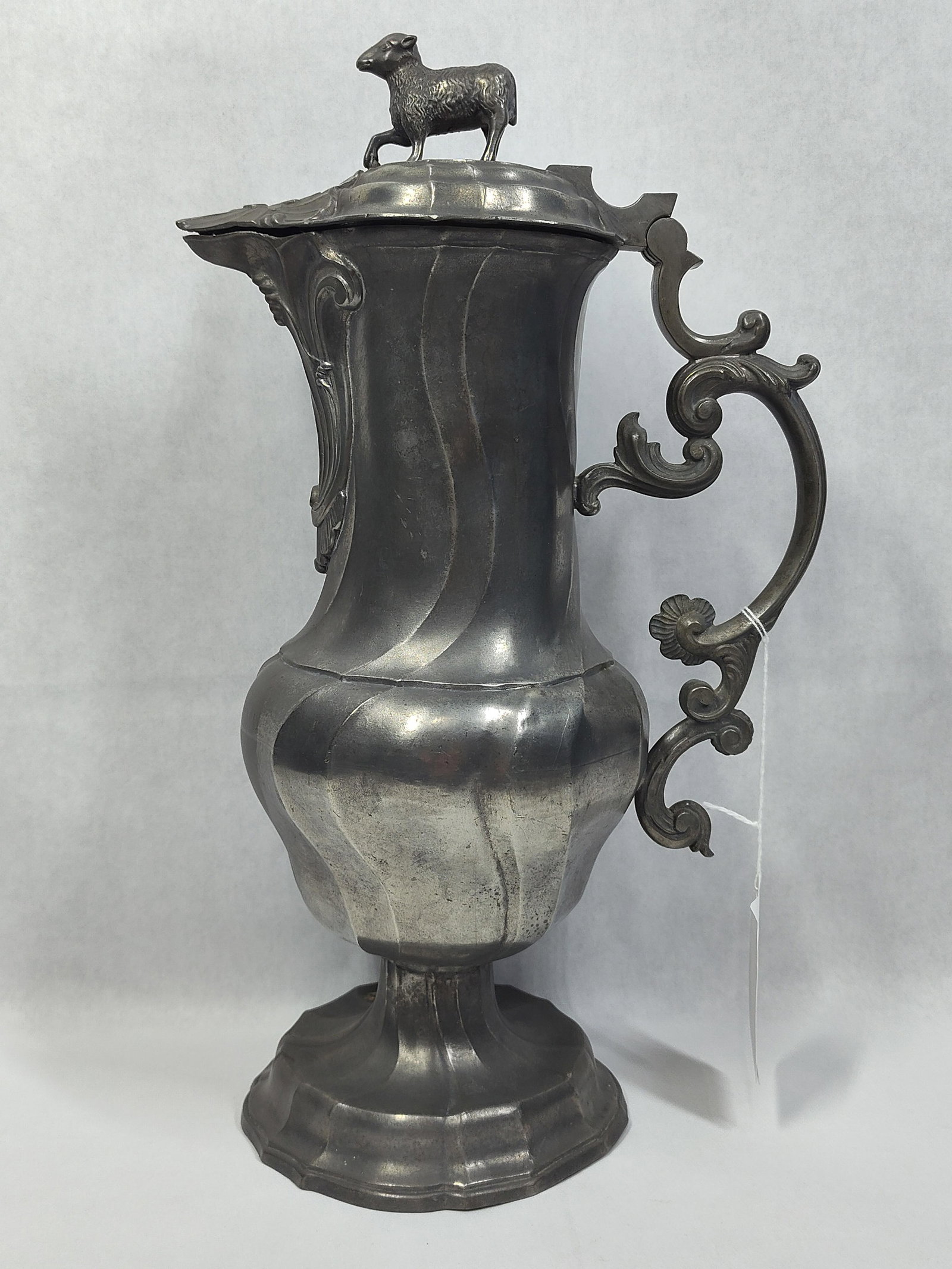 Rococo Style Pewter Flagon with Lamb Finial: This is a pewter flagon or pitcher featuring a hinged lid and elaborate ornamentation in the Rococo style. The vessel has a tall, slender neck transitioning to a bulbous lower body, both decorated