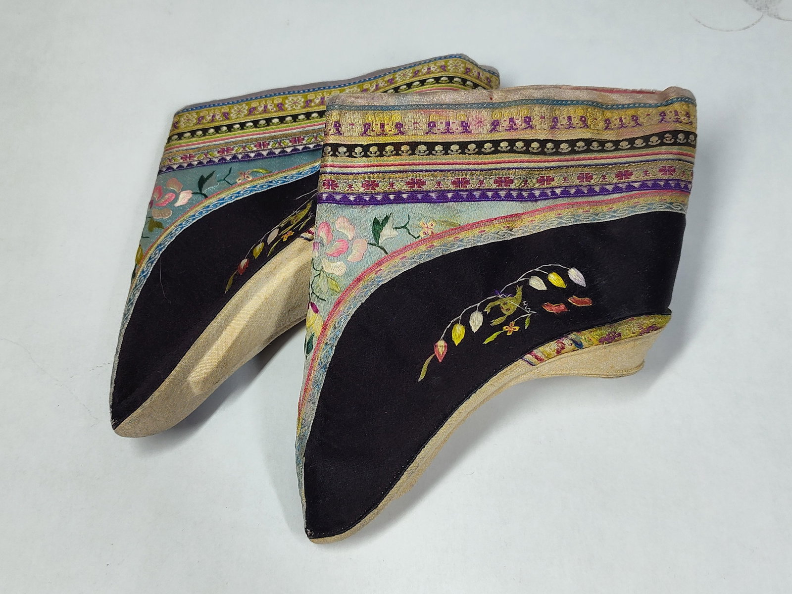 Pair Chinese Embroidered Silk Lotus Shoes, c. 1900: This lot comprises a pair of traditional Chinese lotus shoes intended for women with bound feet, dating from the late 19th to early 20th century. The shoes are constructed with black silk satin uppers