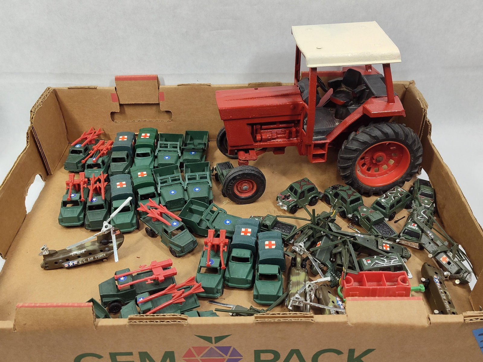 ERTL Int. Harvester Tractor & Military Toys: An assortment of toy vehicles featuring a large red die-cast tractor and various military-themed pieces. The tractor is identified by the ERTL mark embossed on the front tires and is modeled after an