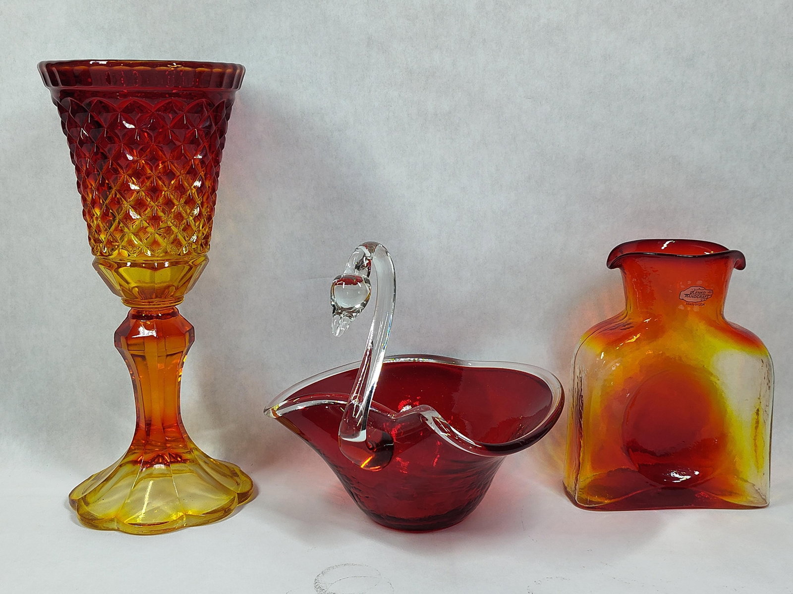 At Glass Yellow to Ruby Red: This collection consists of three pieces of 20th-century American art glass. The first item is a hand-blown glass water carafe by the Blenko Glass Company of Milton, West Virginia. This piece is the