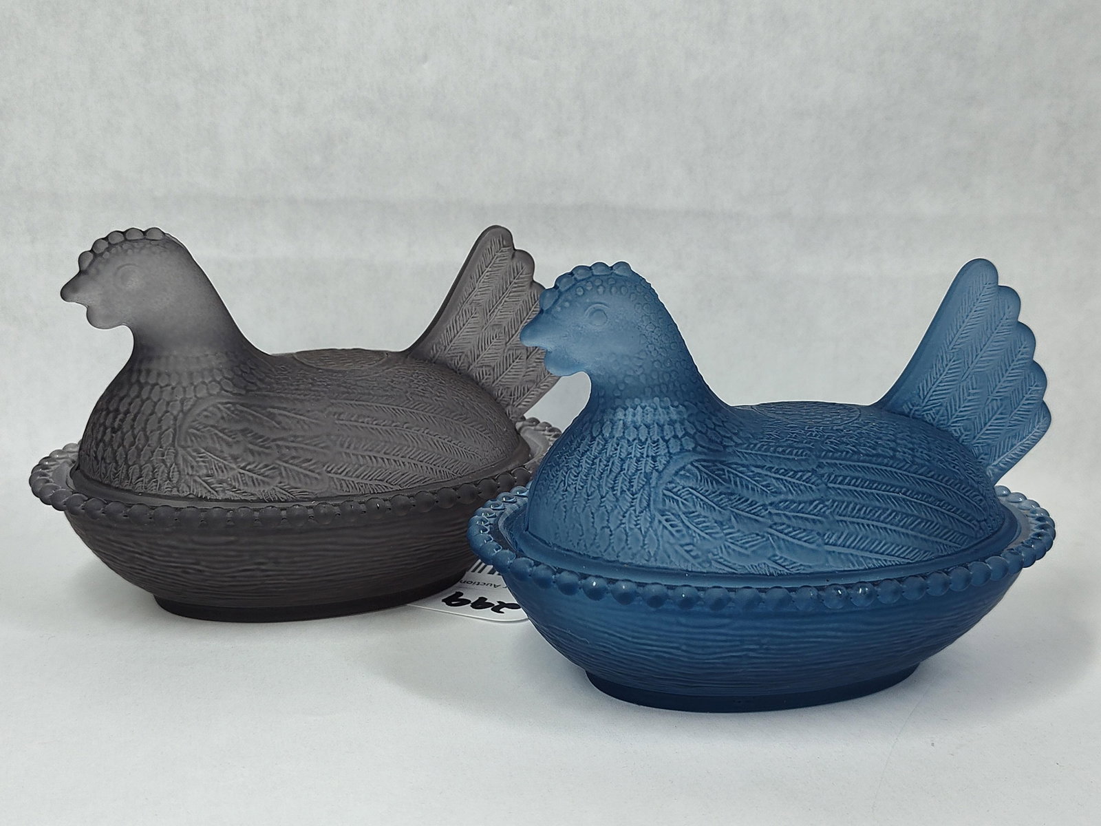 Pair Frosted Satin Glass Hen on Nest Dishes: A pair of matching two-piece covered dishes, each designed in the form of a hen atop a woven basket nest. One dish is rendered in a deep charcoal grey frosted glass, while the other is in a medium