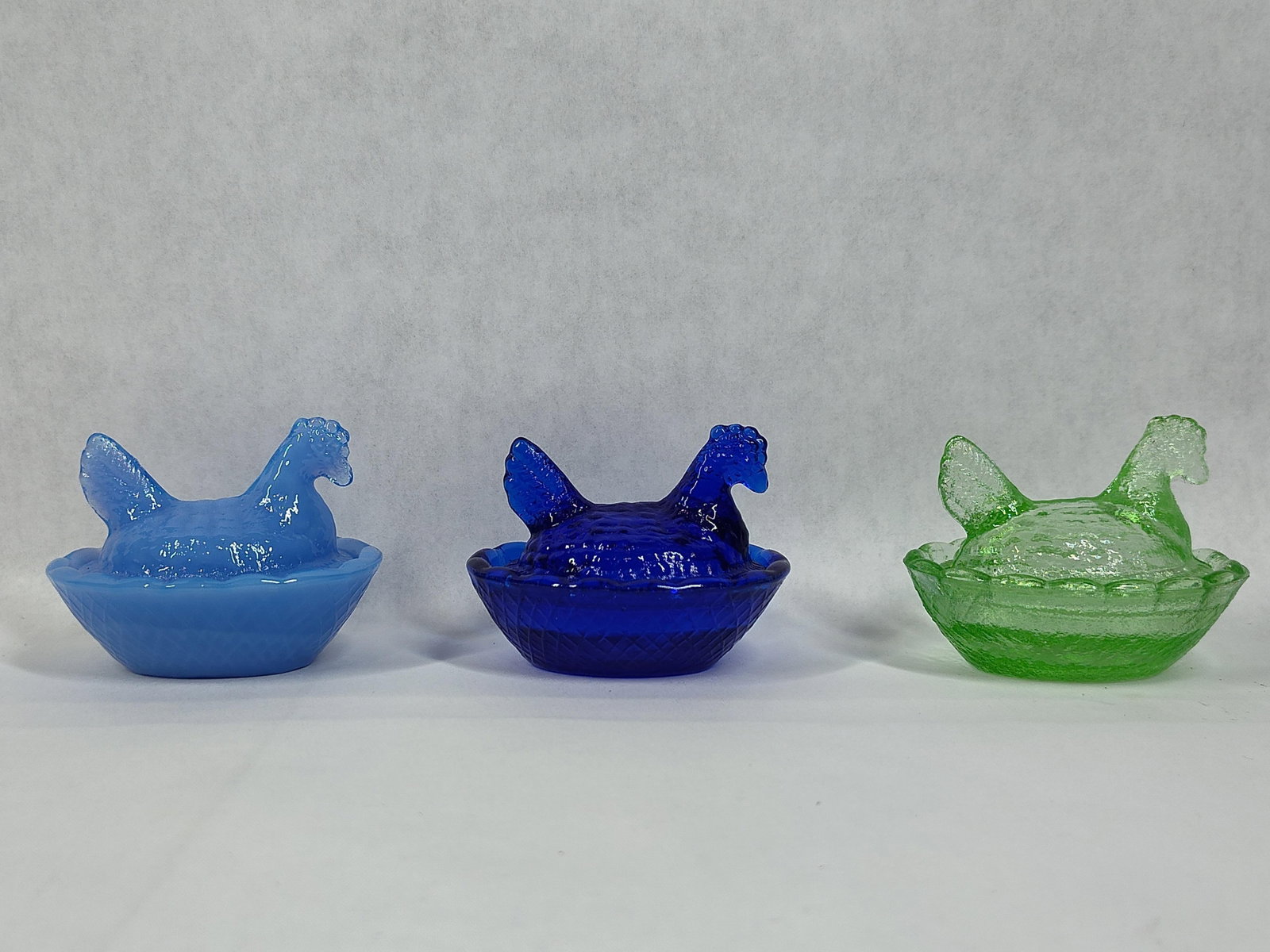 3 Miniature Pressed Glass Hen on Nest Dishes: This lot features a set of three miniature pressed glass lidded dishes designed in the traditional hen on nest form. The assortment includes three different glass types: one in an opaque light blue