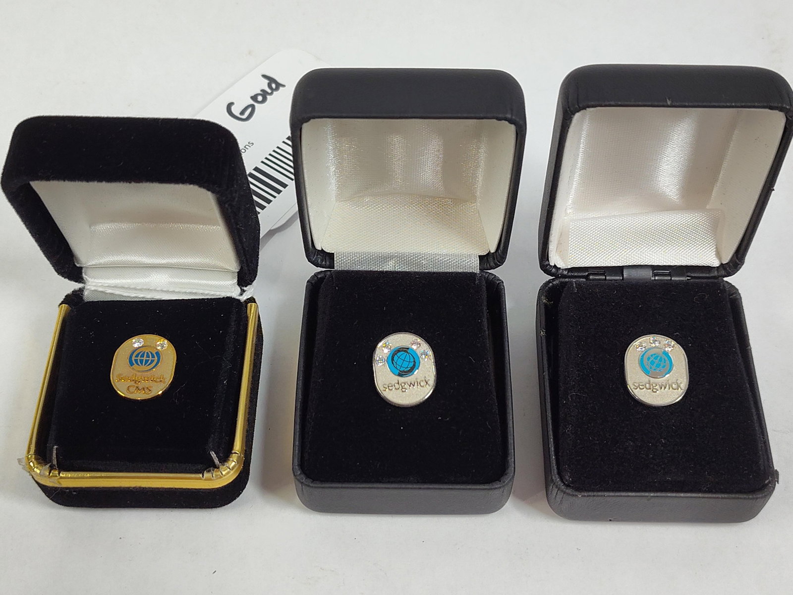 Trio Sedgwick Corporate Service Award Lapel Pins: A trio of corporate service award lapel pins for Sedgwick, each presented in an individual jewelry box. Item 1: One yellow gold-tone oval lapel pin. The face features a central blue globe logo and is