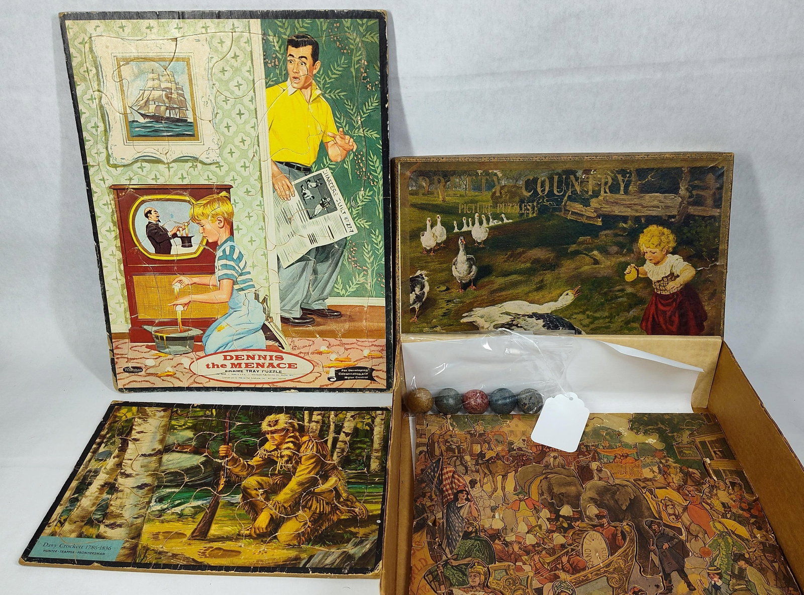 Jigsaw Puzzles & 5 Antique Clay Marbles: This lot features a group of jigsaw puzzles and a set of five antique clay marbles. The puzzle collection includes a Dennis the Menace frame tray puzzle manufactured by the Whitman Publishing Co. of
