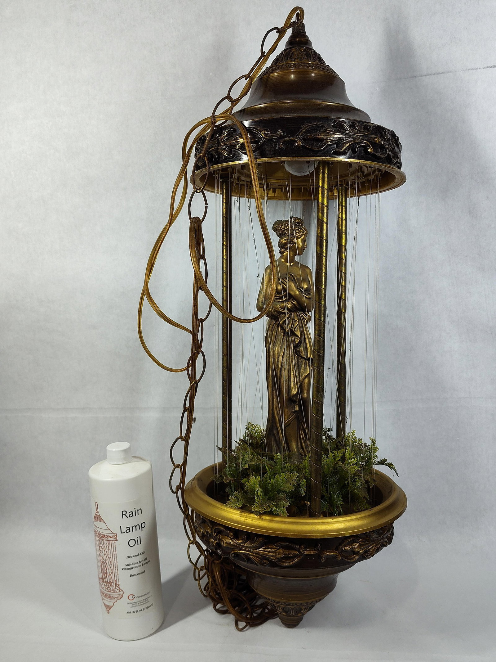 Hanging Gilt Goddess Rain Lamp w/ Faux Foliage: This decorative hanging lamp is designed to circulate mineral oil down monofilament lines to create a visual rain effect. The central feature is a cast classical-style female figure with a metallic