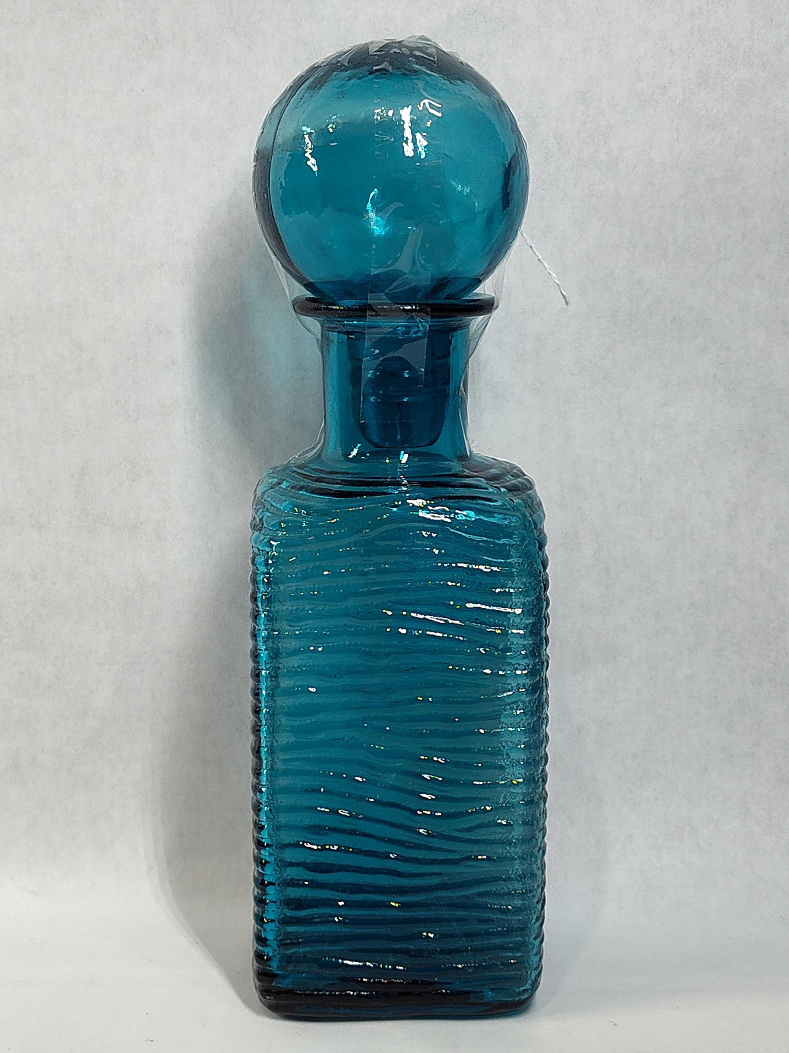 Mid-C. Teal Blue Ripple Glass Decanter w/ Stopper: A Mid-Century Modern molded glass decanter featuring a vibrant teal or turquoise blue color. The vessel is designed with a square-bodied form and a continuous horizontal wavy ripple texture that