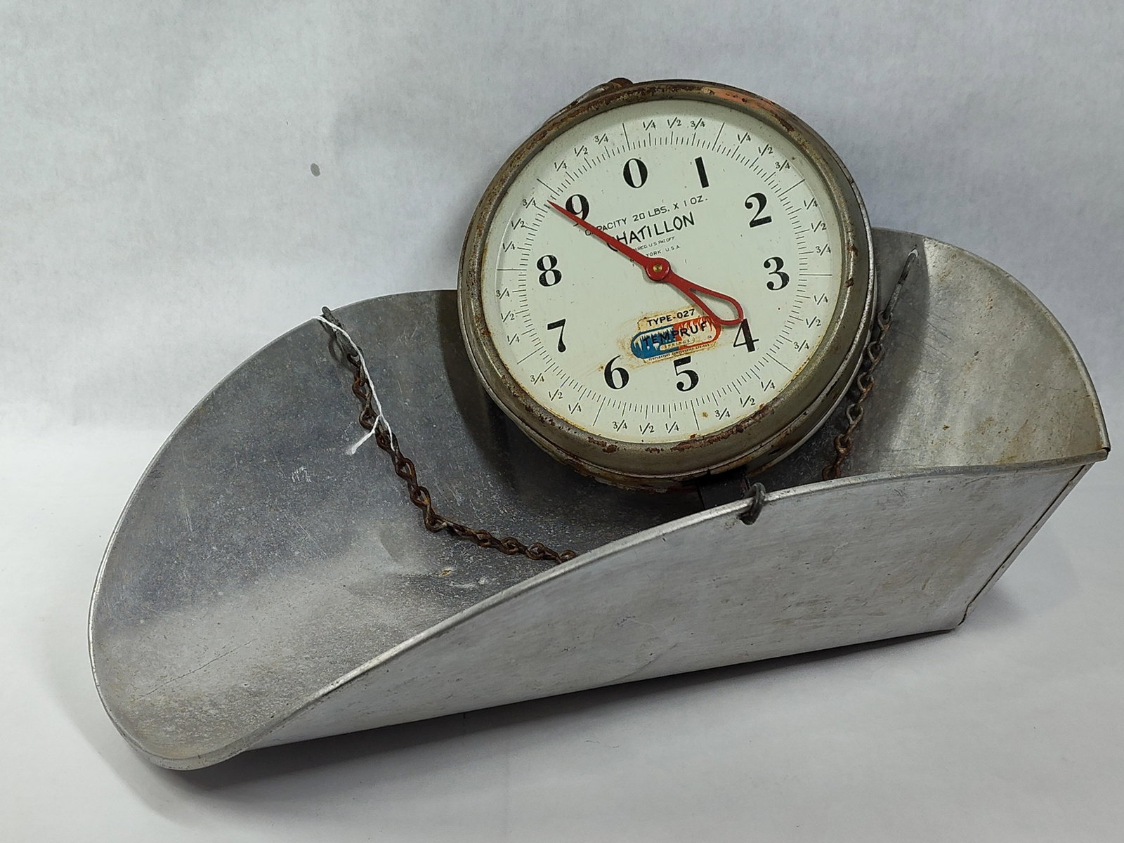 Chatillon Hanging Produce Scale w/ Scoop: This is a Chatillon hanging spring scale, manufactured in New York, USA. The instrument features a circular metal housing with a glass-covered dial face and a red metal indicator needle. The dial is
