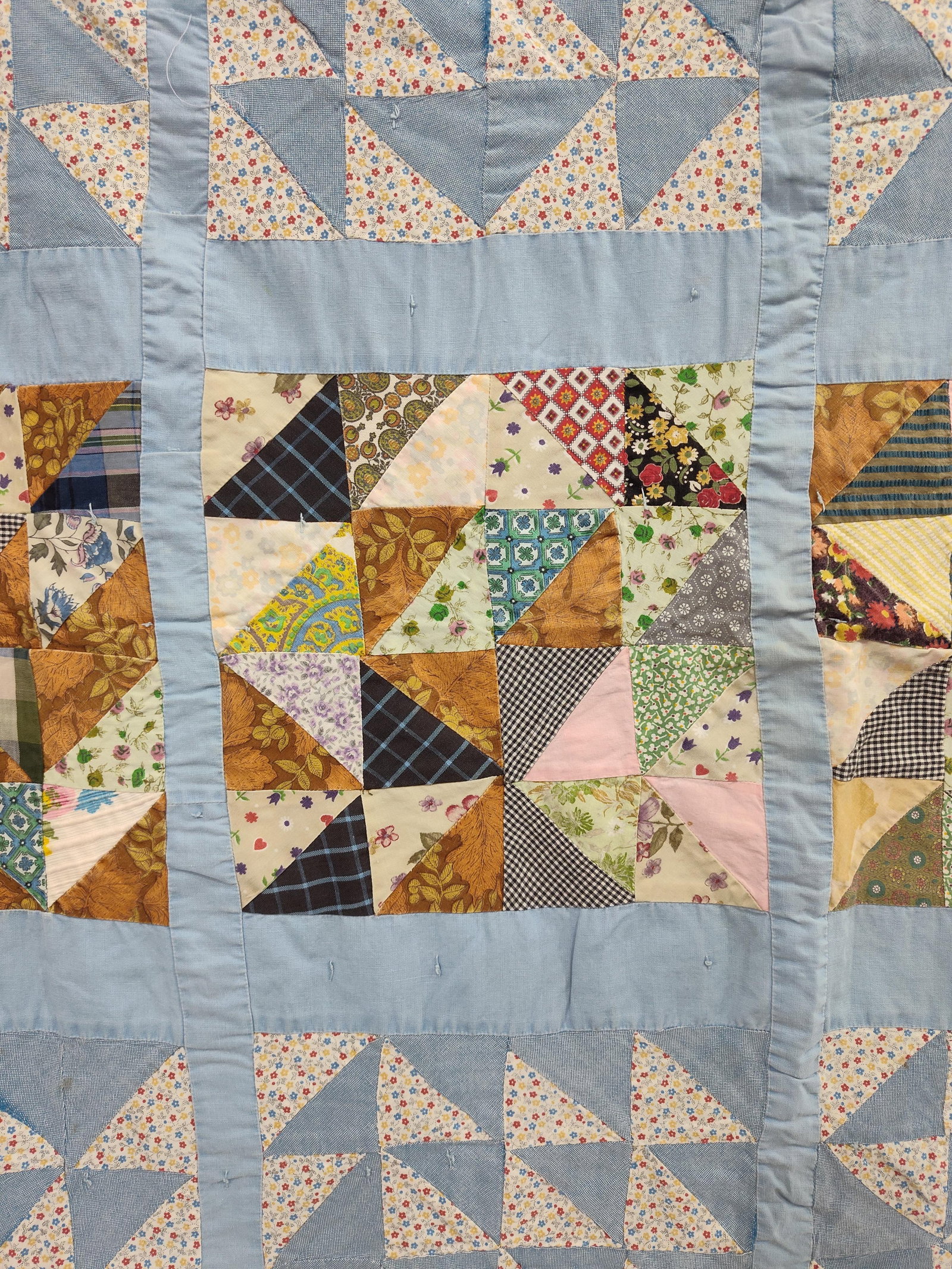American Half-Square Triangle Scrap Quilt: This mid-20th century American utility quilt is hand-pieced and hand-tied. The front features sixteen large blocks arranged in a four-by-four grid, separated by wide light blue cotton sashing. Each