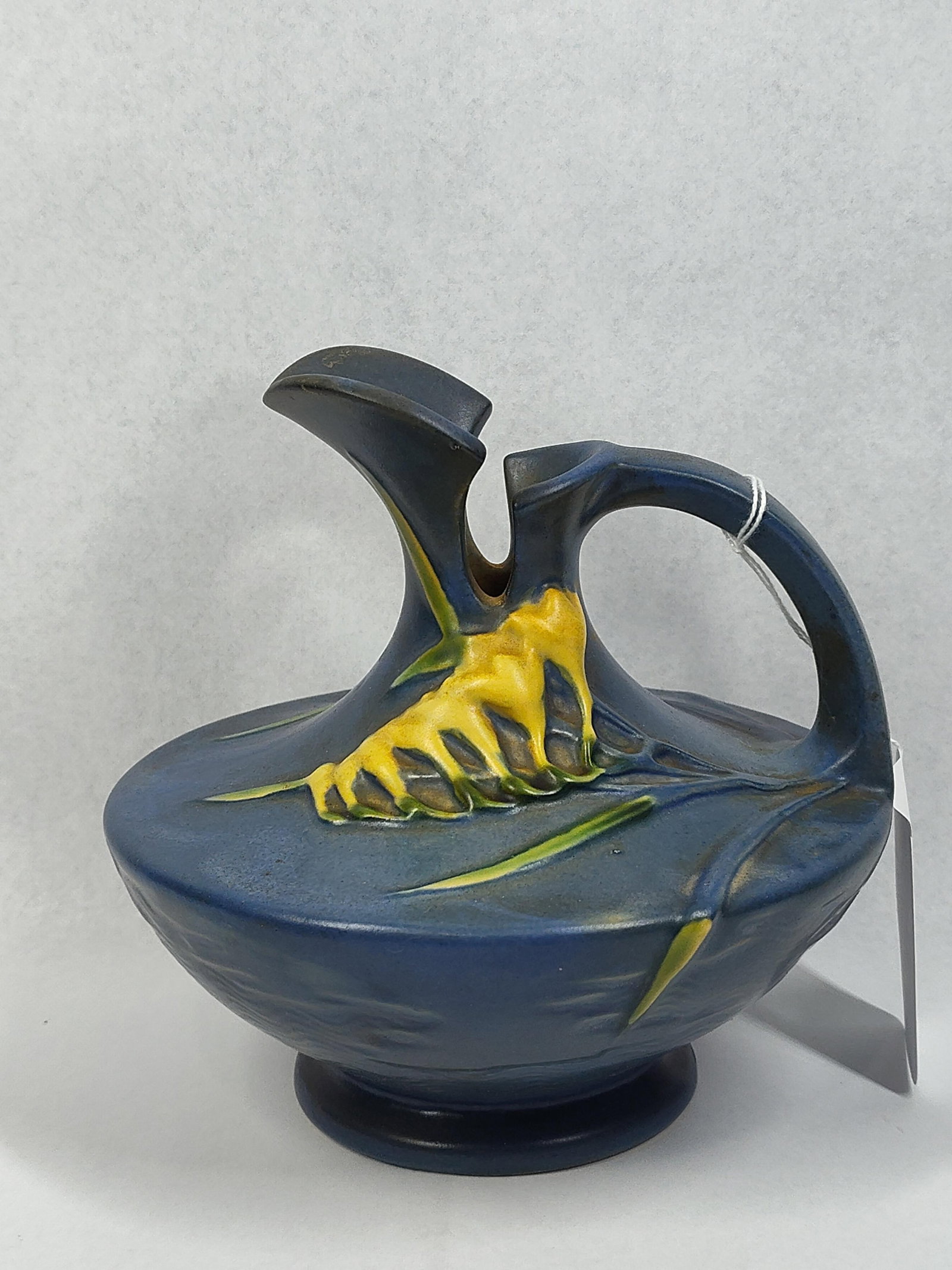 Roseville Pottery Blue Freesia Ewer: 6.5in tall A Roseville Pottery earthenware ewer in the Freesia pattern, originally introduced in 1945. The vessel features a squat, compressed body with a textured matte blue ground. It is decorated w