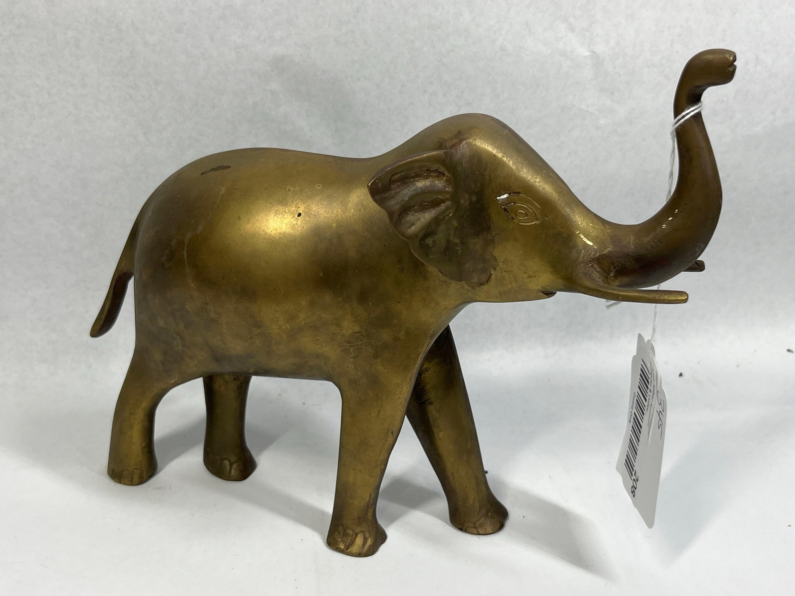 Mid-C. Cast Brass Elephant Raised Trunk: 11in A mid-century style decorative figurine of an elephant crafted from cast brass. The figure is depicted in a walking pose with its trunk raised in a traditional gesture of good luck. It features