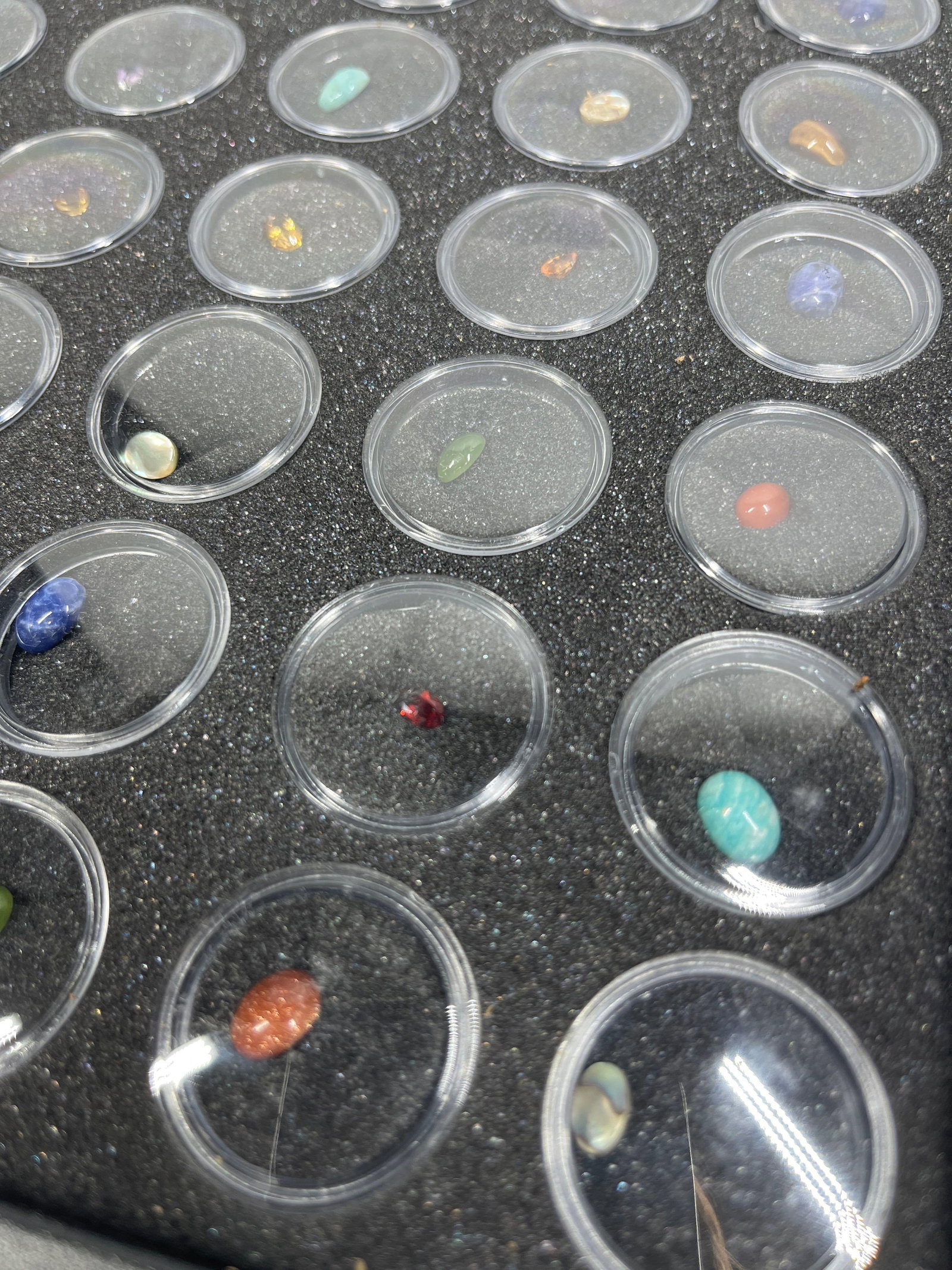 100 Mixed Loose Gemstones w/ Trays: This collection consists of 100 individual loose gemstones, professionally housed in clear plastic circular jars. The collection is organized into two black foam-lined display trays, each containing