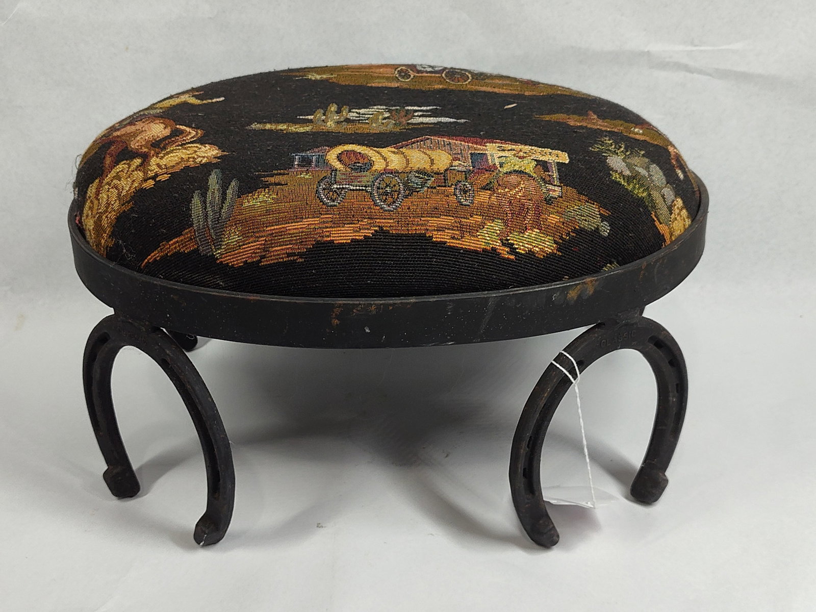 Western Horseshoe Footstool w/ Tapestry: 15x12x8 This small footstool features an oval seat upholstered in a woven tapestry fabric. The decorative fabric displays a repeating Western frontier motif against a black background, including a