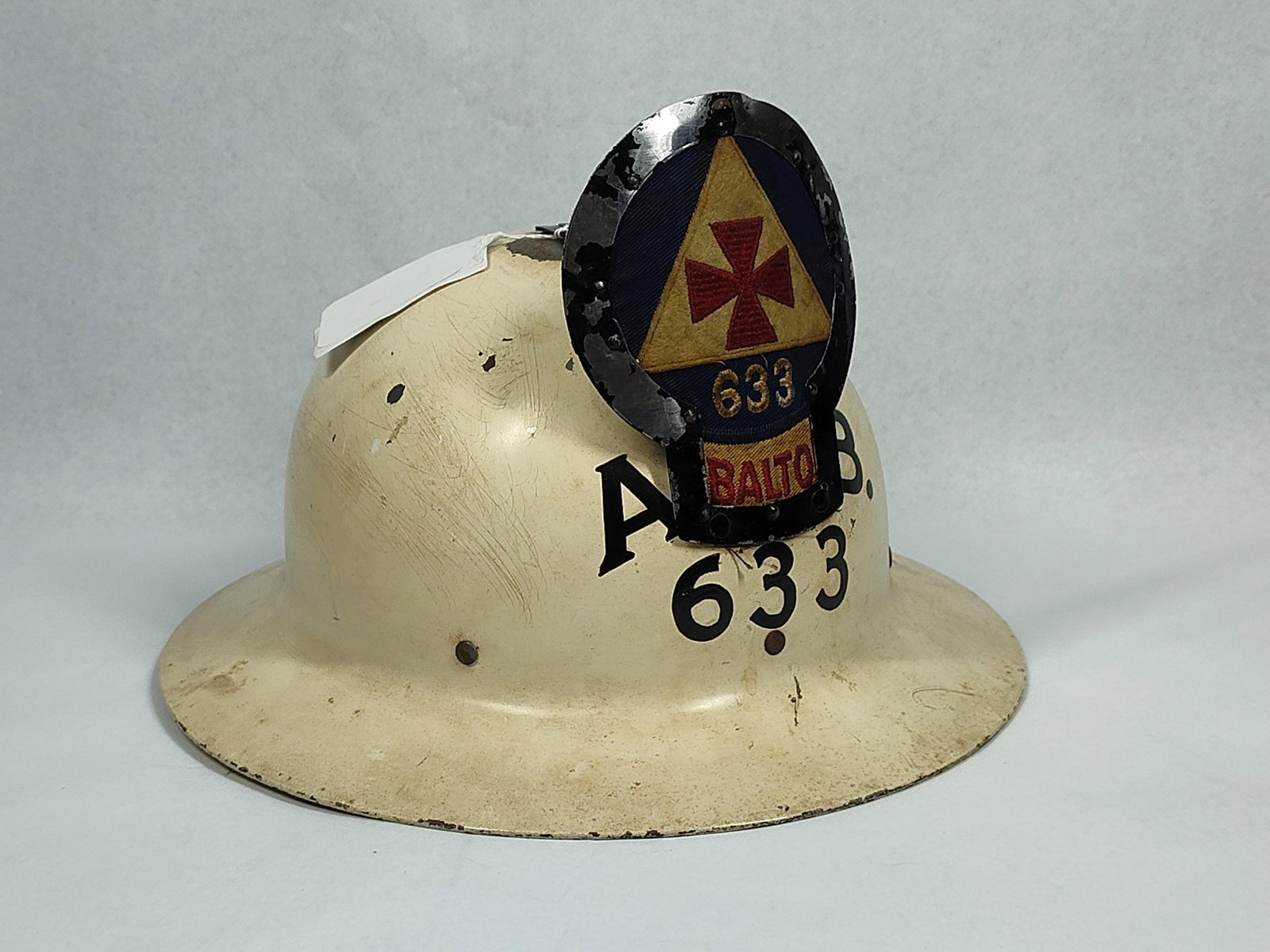 WWII US OCD Baltimore Red Cross Helmet: A WWII-era United States Office of Civilian Defense steel helmet. The object is a Brodie-style steel helmet finished in white paint. A black-painted metal identification bracket is bolted to the
