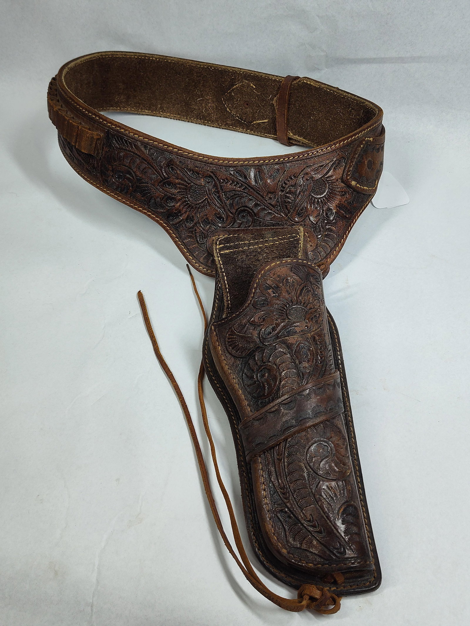 Hand-Tooled Leather Buscadero Rig: A Western-style buscadero gun belt and matching drop-loop holster constructed from dark brown leather. The rig features extensive hand-tooling in a floral and scrollwork pattern across the entirety