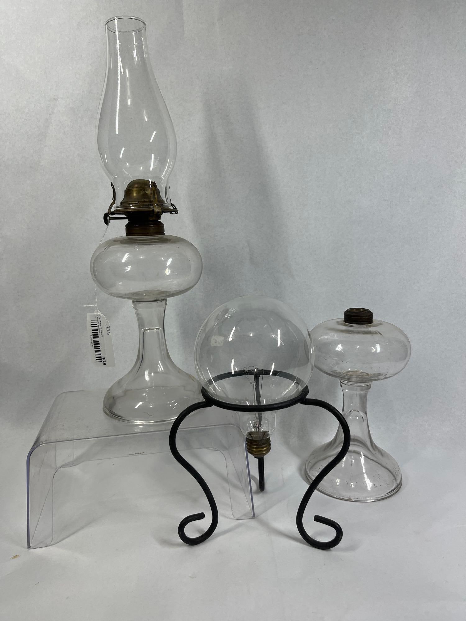 3 Glass Pedestal Oil Lamps & Bulb Stand: This lot features three lighting-related decorative items. The collection includes one complete kerosene table lamp constructed from clear pressed glass with a pedestal base and rounded font. It is