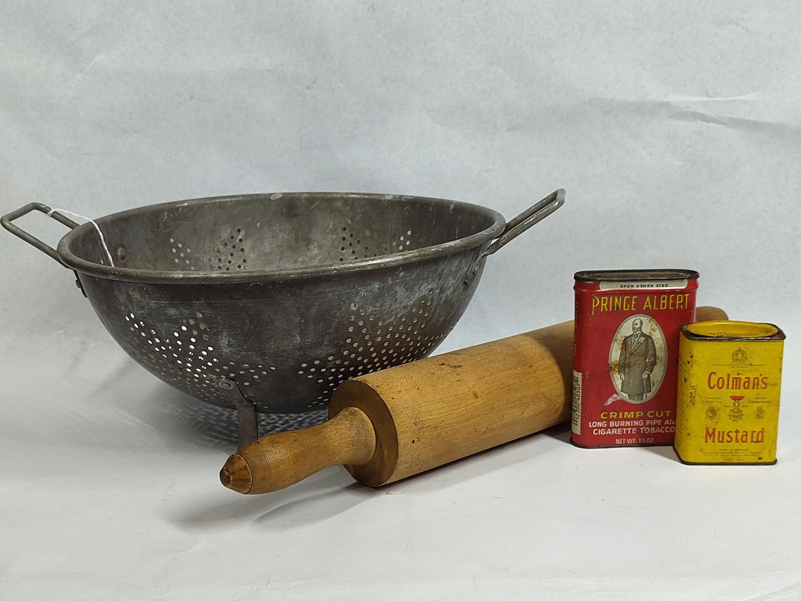 Kitchenware & Advertising Tins: An assortment of vintage household items featuring a footed metal colander, a wooden rolling pin, and two lithographed advertising tins. The colander, resembling designs by Wear-Ever, is crafted from
