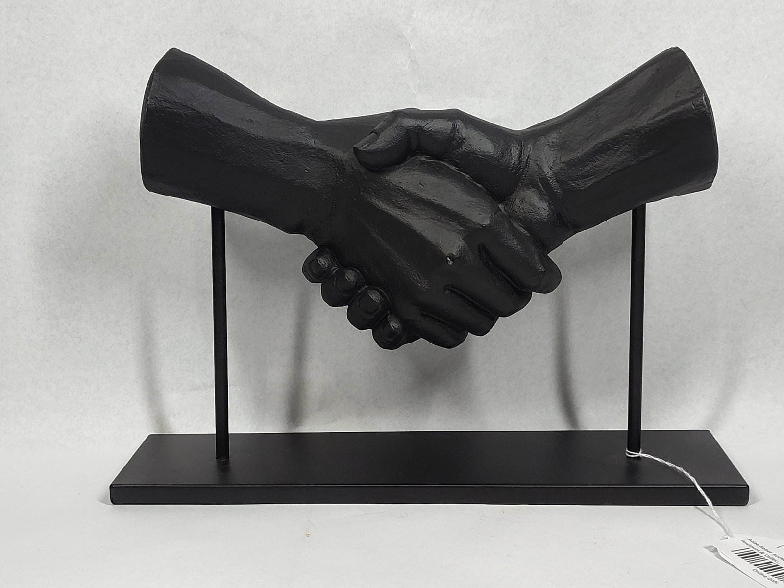 Handshake Sculpture on Stand: The piece depicts two hands joined in a handshake, made with plastic or a lightweight material, finished in a uniform matte black coating. The sculpture is elevated on two slender vertical metal rods