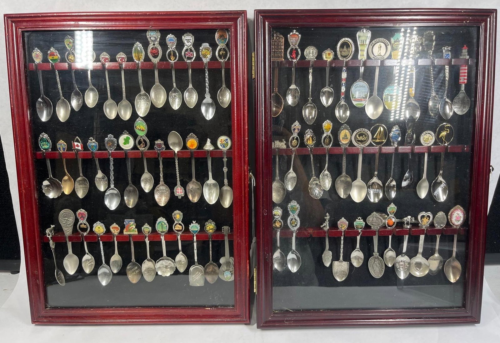 Souvenir Spoons in 2 Wood Display Cases: 19x14 An assortment of souvenir spoons presented in two wall-mounted wooden display cases. The cases are finished in a red tone with glass fronts and black felt-lined interiors featuring horizontal