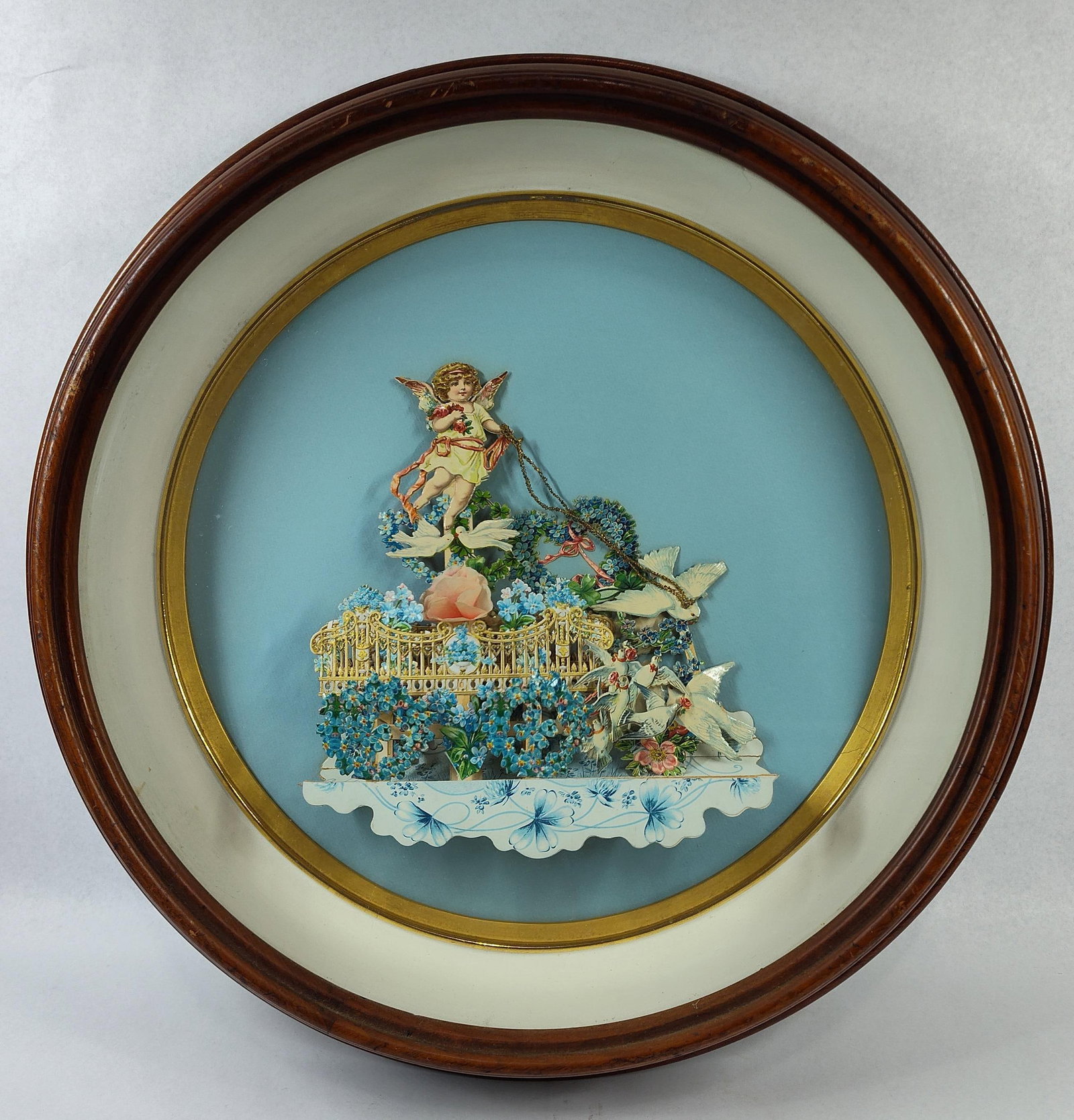Victorian 3D Chromolithograph Diorama: This lot consists of a circular shadowbox frame containing a three-dimensional chromolithographed paper diorama, resembling a Victorian-era German mechanical pull-down valentine. The central assembly