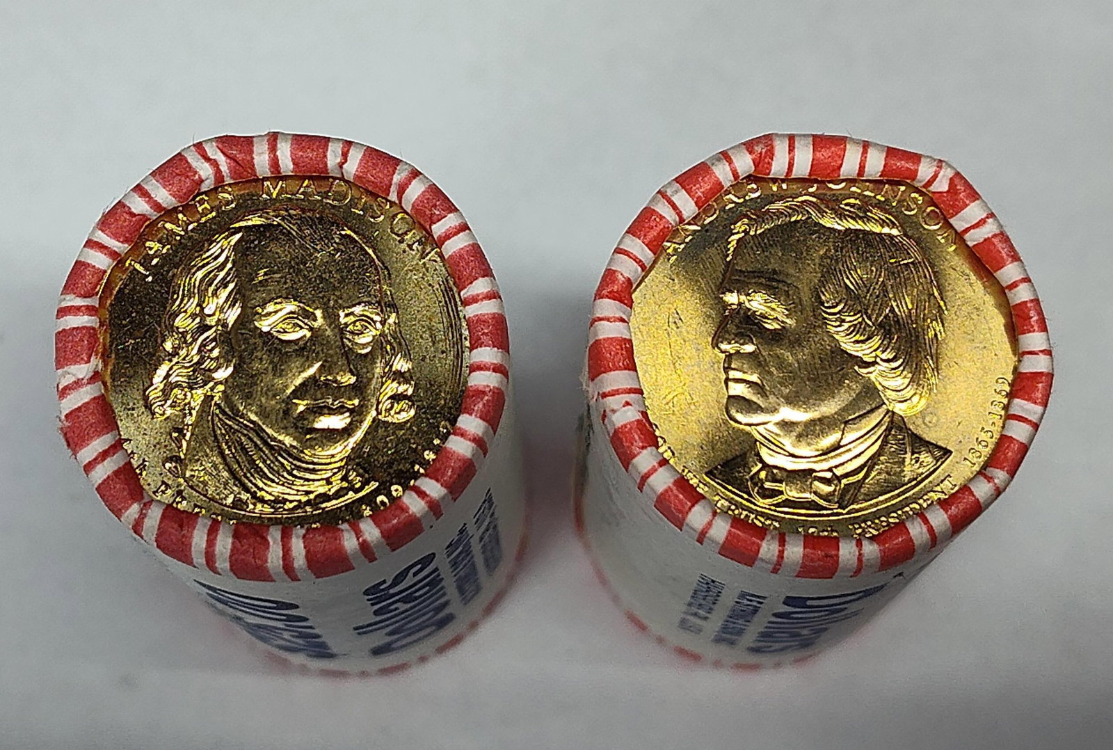 Pair Madison & Johnson US $1 Coin Rolls: A pair of bank-wrapped rolls containing US Presidential $1 coins, specifically the James Madison (2007) and Andrew Johnson (2011) issues. Each roll comprises twenty-five manganese brass-clad copper