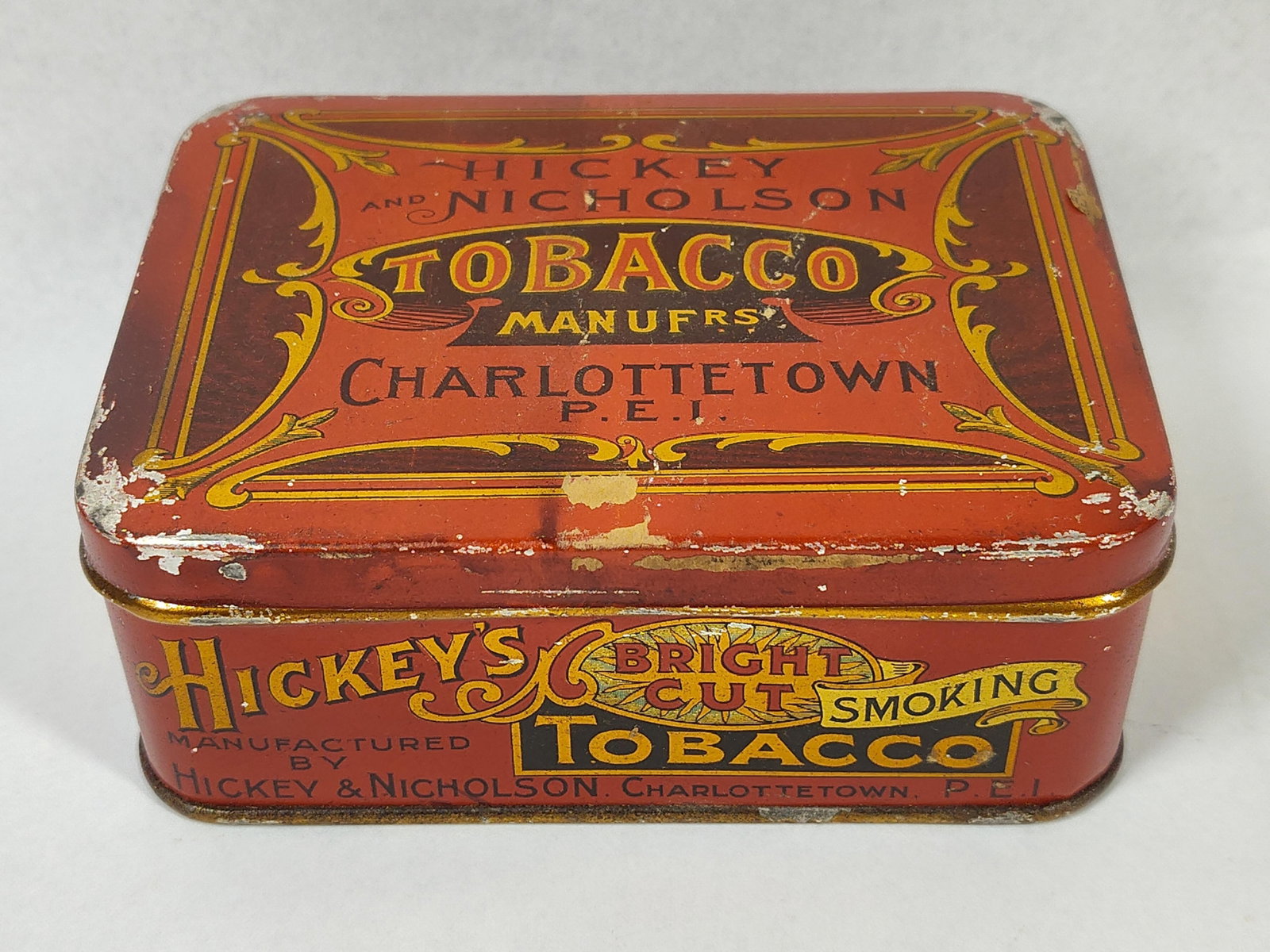 Hickey & Nicholson Tobacco Tin & Pipe: This lot features a rectangular lithographed pocket tobacco tin and a miniature novelty smoking pipe. The tin is finished in a red background with ornate gold and black typography and scrollwork. The