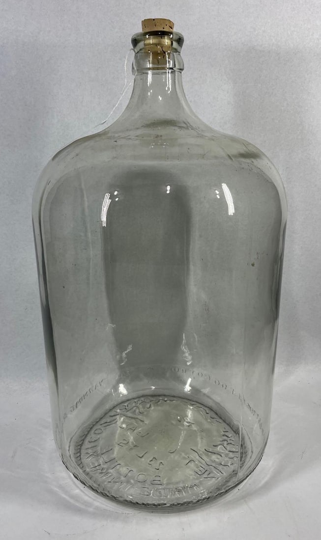 Vitro Mexico Eagle Emblem Glass Carboy: 20x10. A large 6-gallon clear glass carboy manufactured by Vitro in Mexico. The vessel features a cylindrical body with a tapered neck and a reinforced rim, currently fitted with a tapered cork
