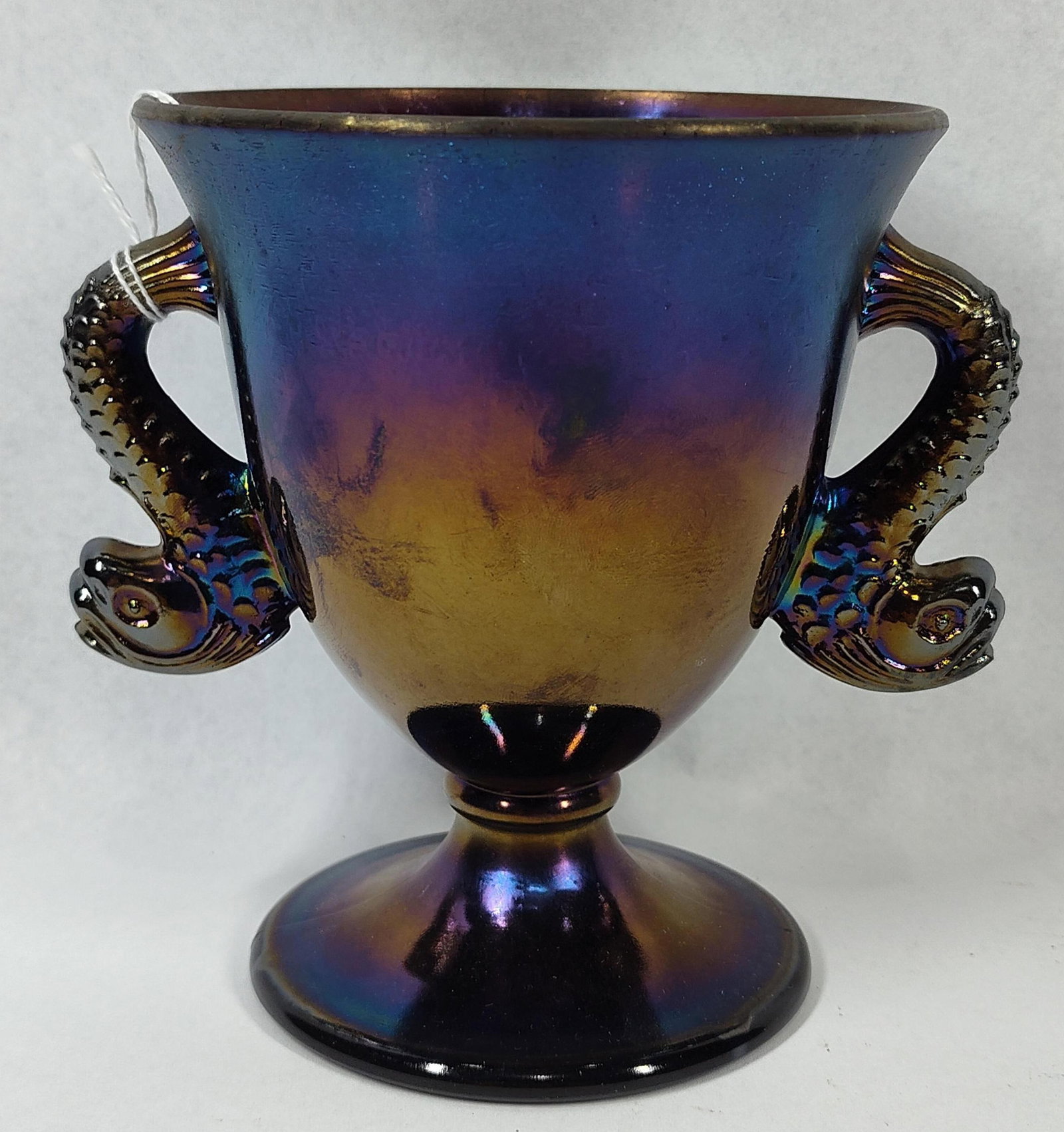 Amethyst Carnival Dolphin Loving Cup: 5.5 in tall. This pressed carnival glass vessel is a two-handled loving cup or urn, attributed to the Dolphin pattern. The piece features a tapered cup body with a flared rim, supported by a short