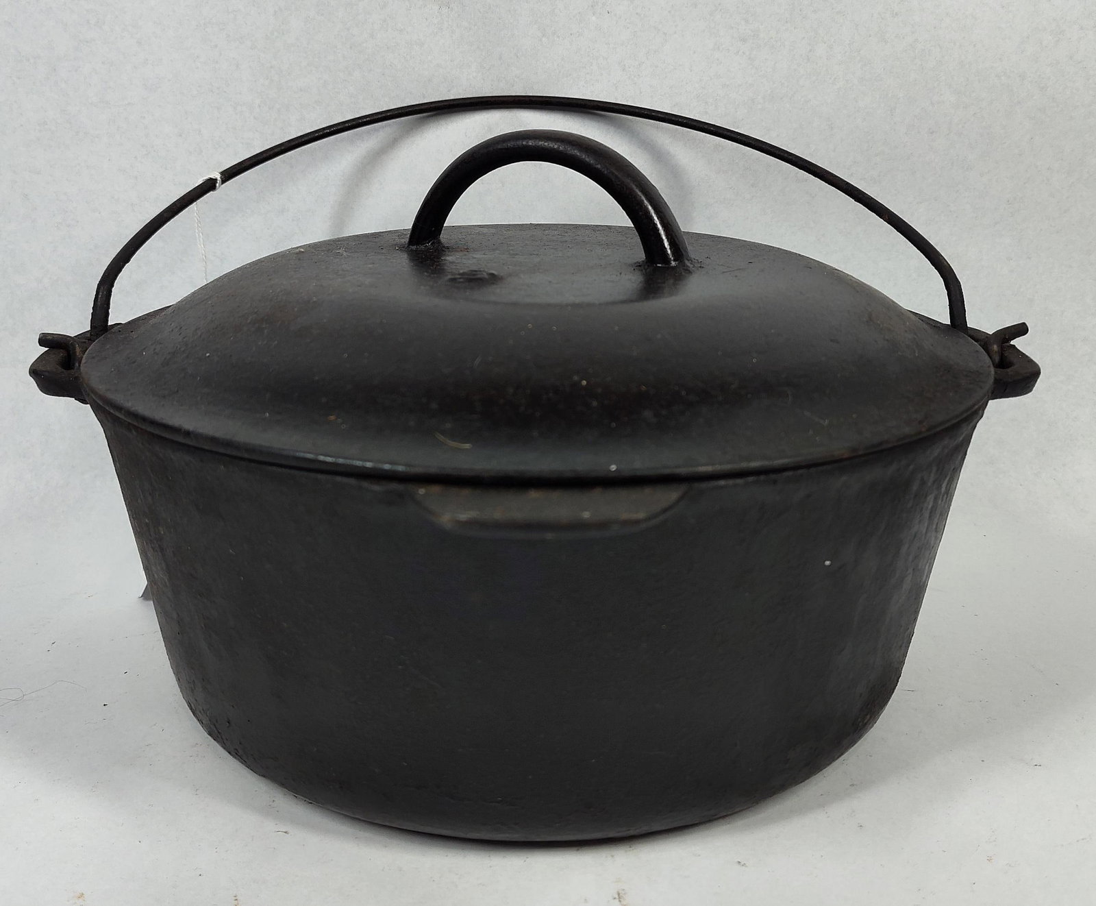 Vtg Cast Iron Dutch Oven w/ Basting Lid: A vintage cast iron Dutch oven comprising a round-bodied pot, a matching domed lid, and a wire bail handle. The pot is designed with a flat bottom and two integral side ears for handle attachment. The