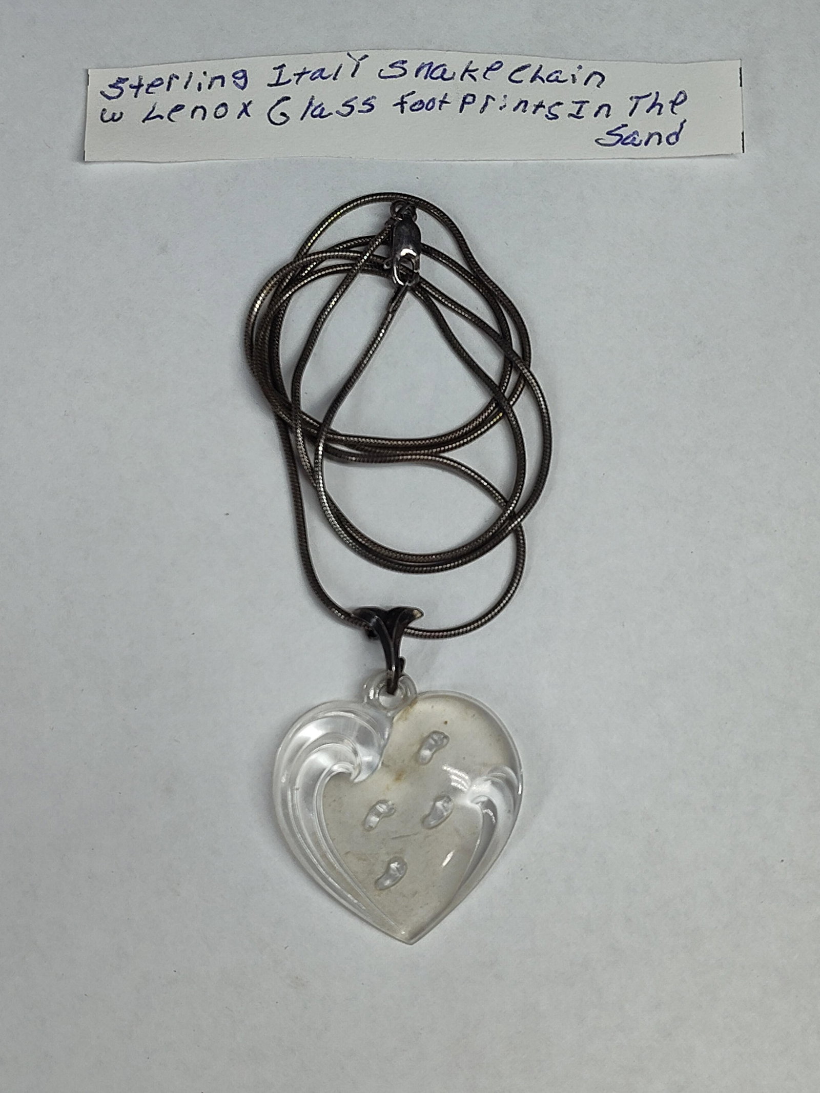 Lenox Footprints Glass Heart Str. Chain: This necklace features a clear glass heart-shaped pendant suspended from a sterling silver snake chain. Attributed to Lenox and featuring the Footprints in the Sand motif, the pendant contains four
