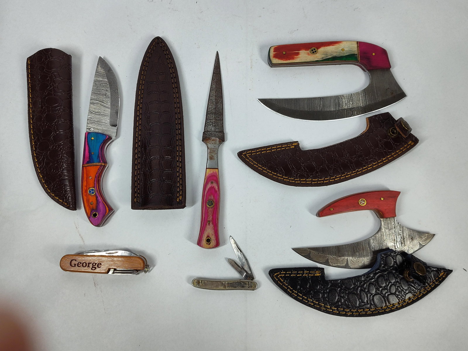 Damascus Steel Fixed & Folding Knives: This collection consists of several knives, including multiple fixed-blade pieces made from pattern-welded Damascus steel. The assortment features a hunter-style knife with a laminated wood handle in