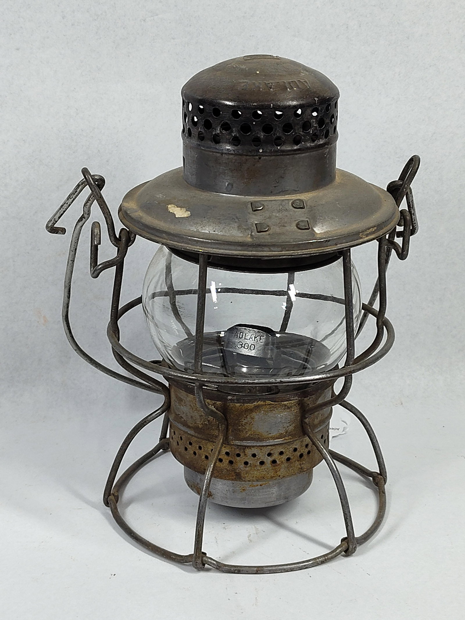 Adlake Kero No. 300 Railroad Lantern: This is an Adlake Kero No. 300 railroad lantern manufactured by the Adams & Westlake Company. The lantern features a steel wire cage frame designed to protect a bulbous clear glass globe. The