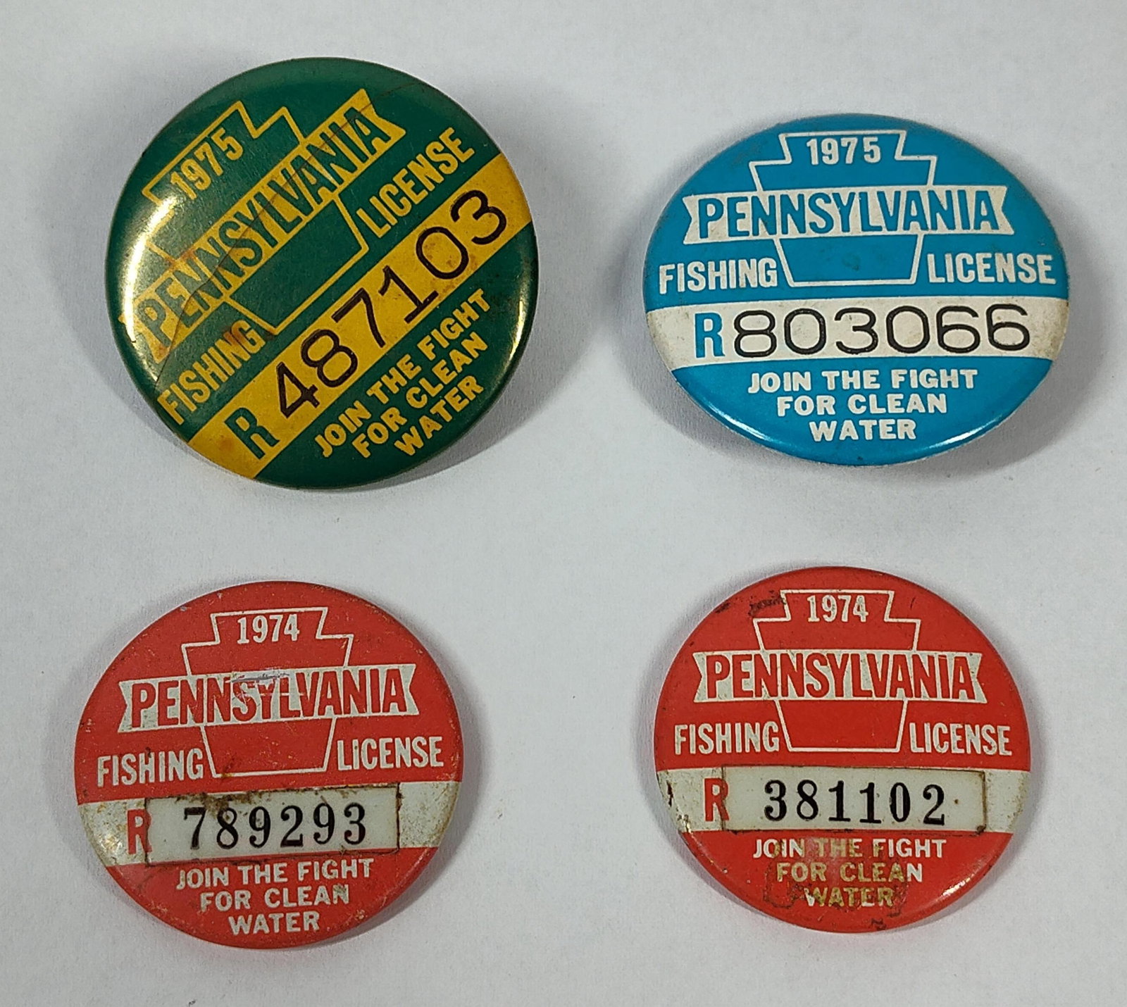 4 Vtg PA Fishing License Buttons '74-75: A collection of four vintage metal pinback fishing licenses issued by the state of Pennsylvania. This lot contains two licenses from 1974 and two from 1975. The 1974 licenses are designed in red and
