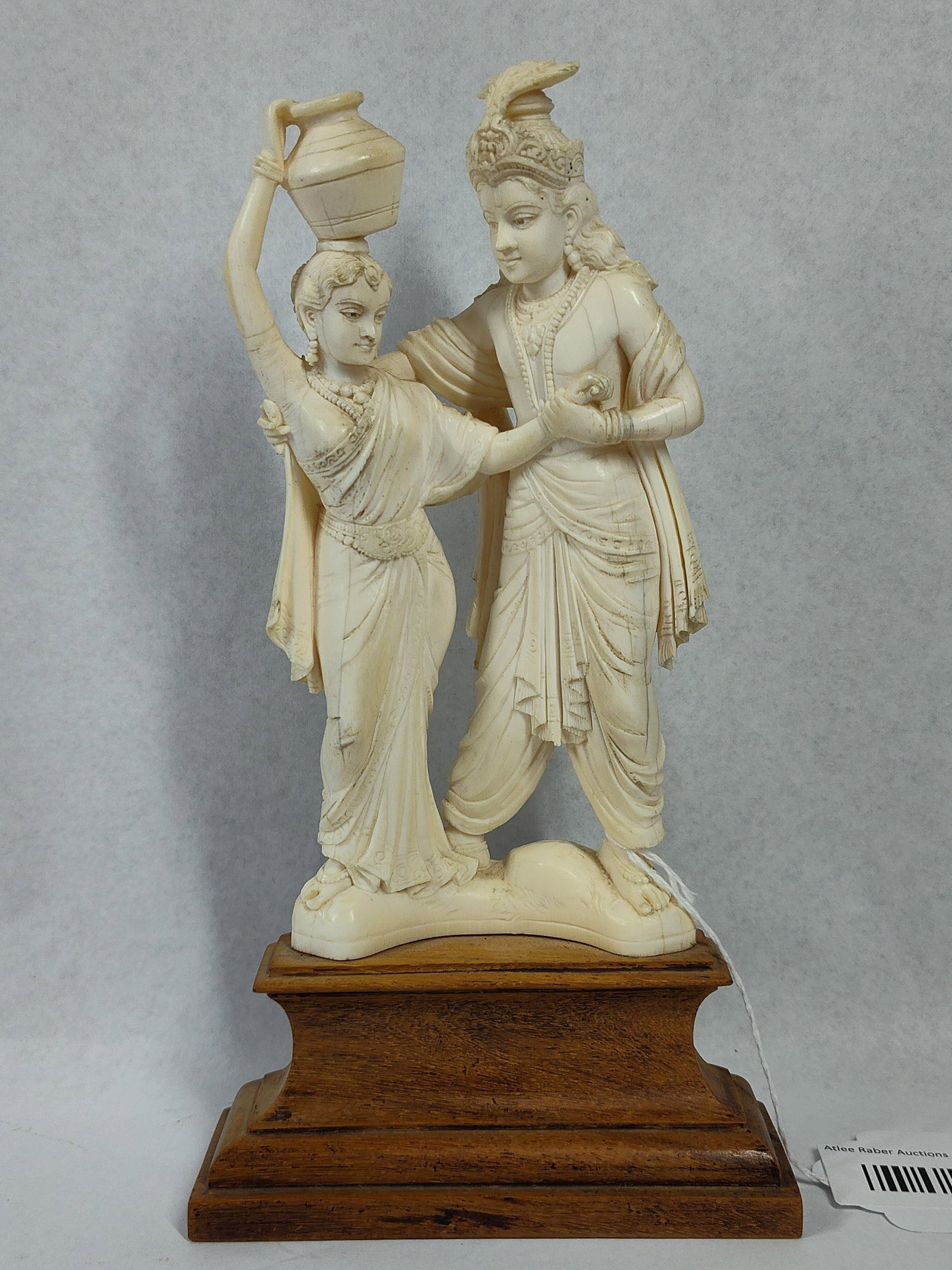 Indian Wood Radha-Krishna Figural Group: 9 inches tall. A carved figural sculpture depicting the Hindu deities Radha and Krishna, mounted on a tiered wooden base. The figures are rendered in a traditional style from an off-white, cream-toned