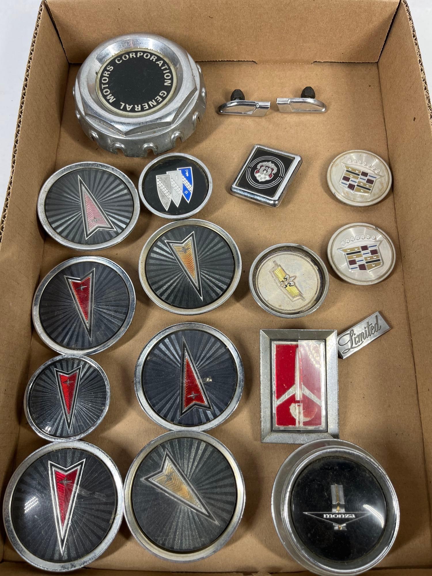 Vtg General Motors Wheel Caps & Emblems: This collection consists of various automotive components representative of General Motors divisions from the 1970s and 1980s. The assortment includes several circular wheel center caps featuring the