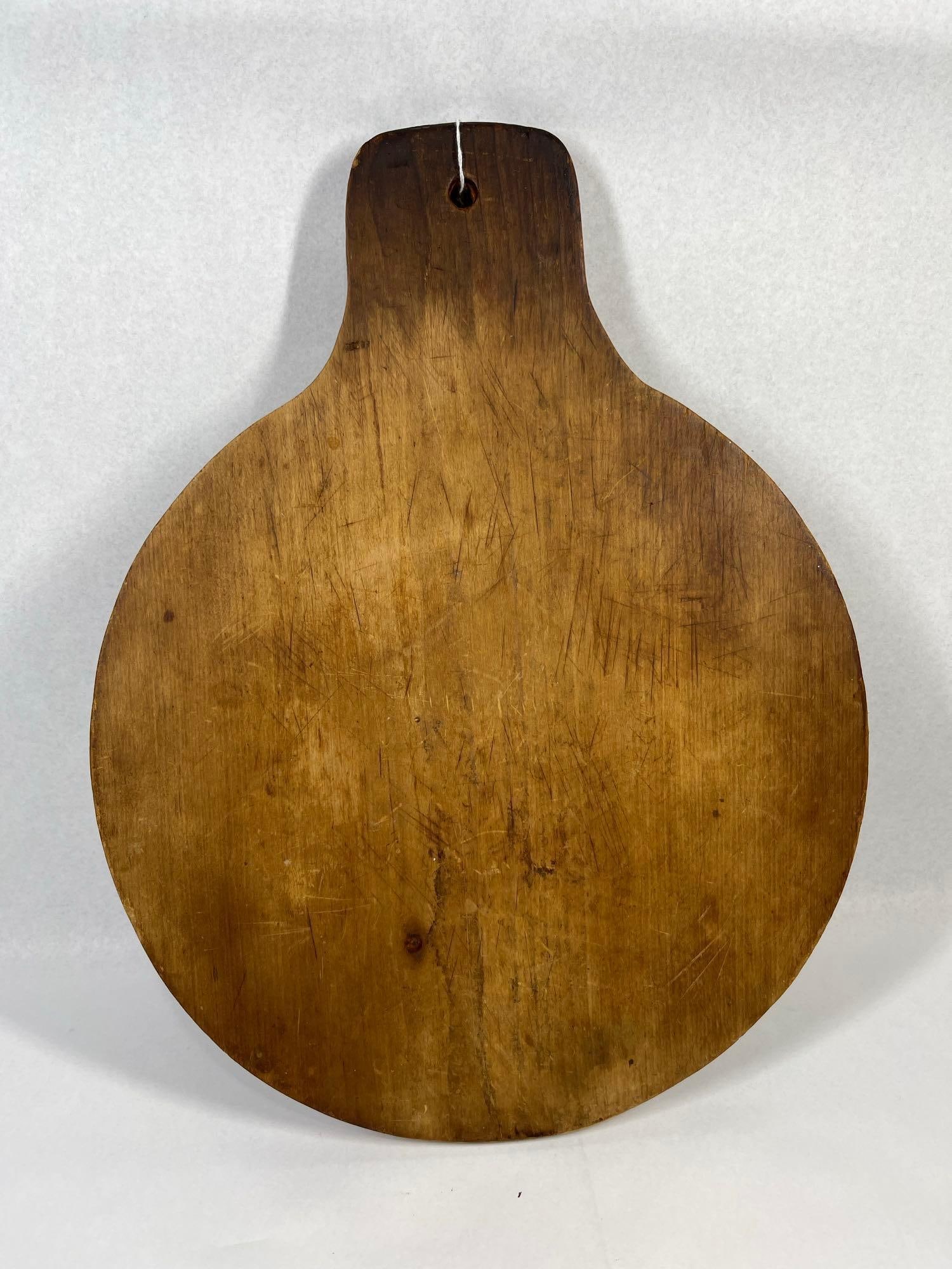Primitive Round Cutting Board w/ Handle: 19.5x15 This primitive kitchen tool is a circular cutting board or bread board crafted from a single piece of hardwood. The board features a wide, rounded body that tapers into a short, integral