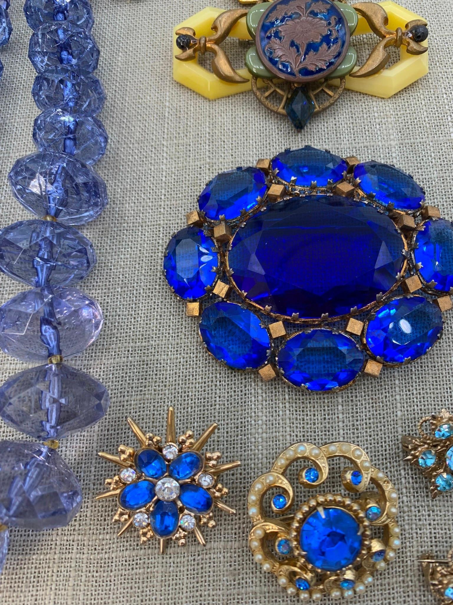 Bourjois Vial & Art Deco Blue Jewelry: This collection consists of an assortment of vintage costume jewelry and vanity items primarily featuring blue glass and rhinestone accents. A prominent piece is a large gold-tone cluster brooch