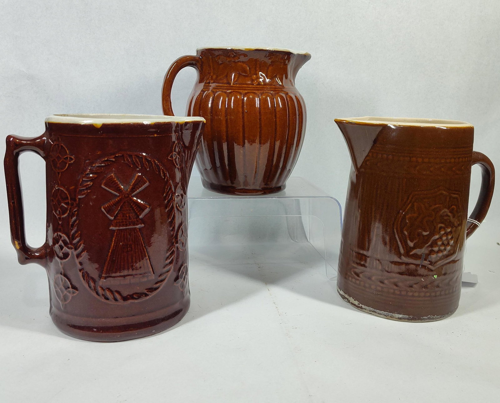 3 Sea Captain Relief Stoneware Pitchers: 7.25 to 8 in. A group of three relief-molded stoneware pitchers featuring dark brown glazes with contrasting white or cream-glazed interiors. One pitcher is attributed to Burley Winter Pottery of